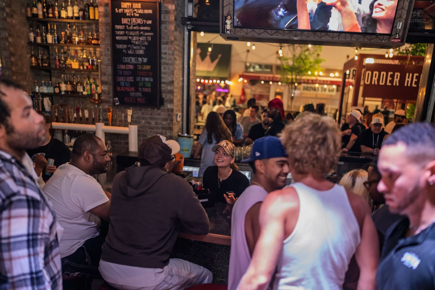 Diverse group of people socializing at a busy indoor-outdoor bar venue with brick walls and warm ambient lighting.
