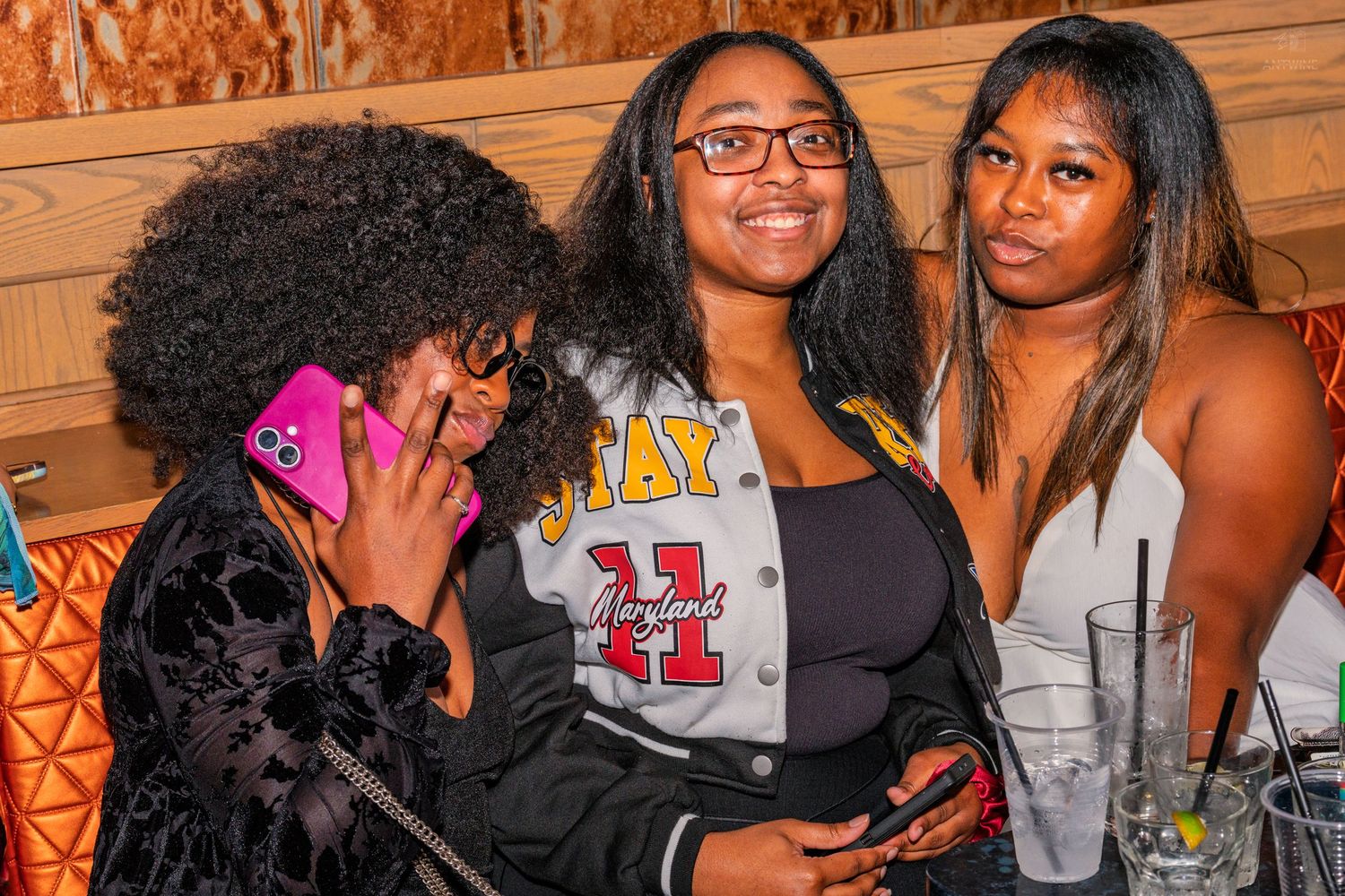 Three women smiling together at a bar, one holding a pink phone.