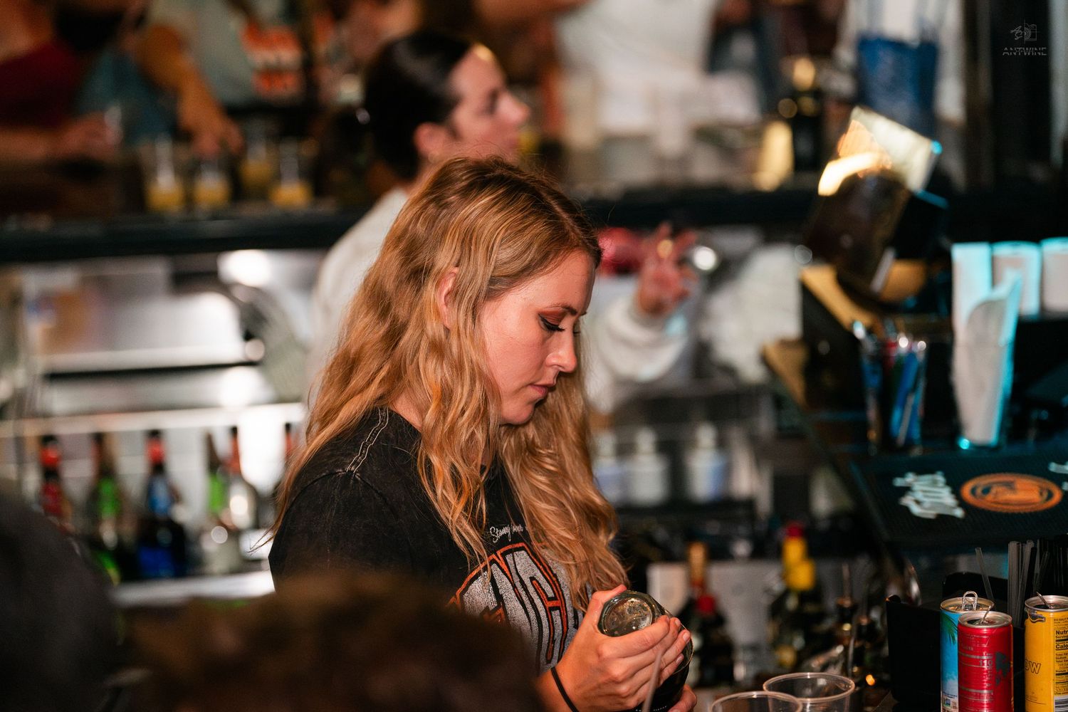 Female bartender carefully preparing a cocktail at a busy bar with bottles in background.