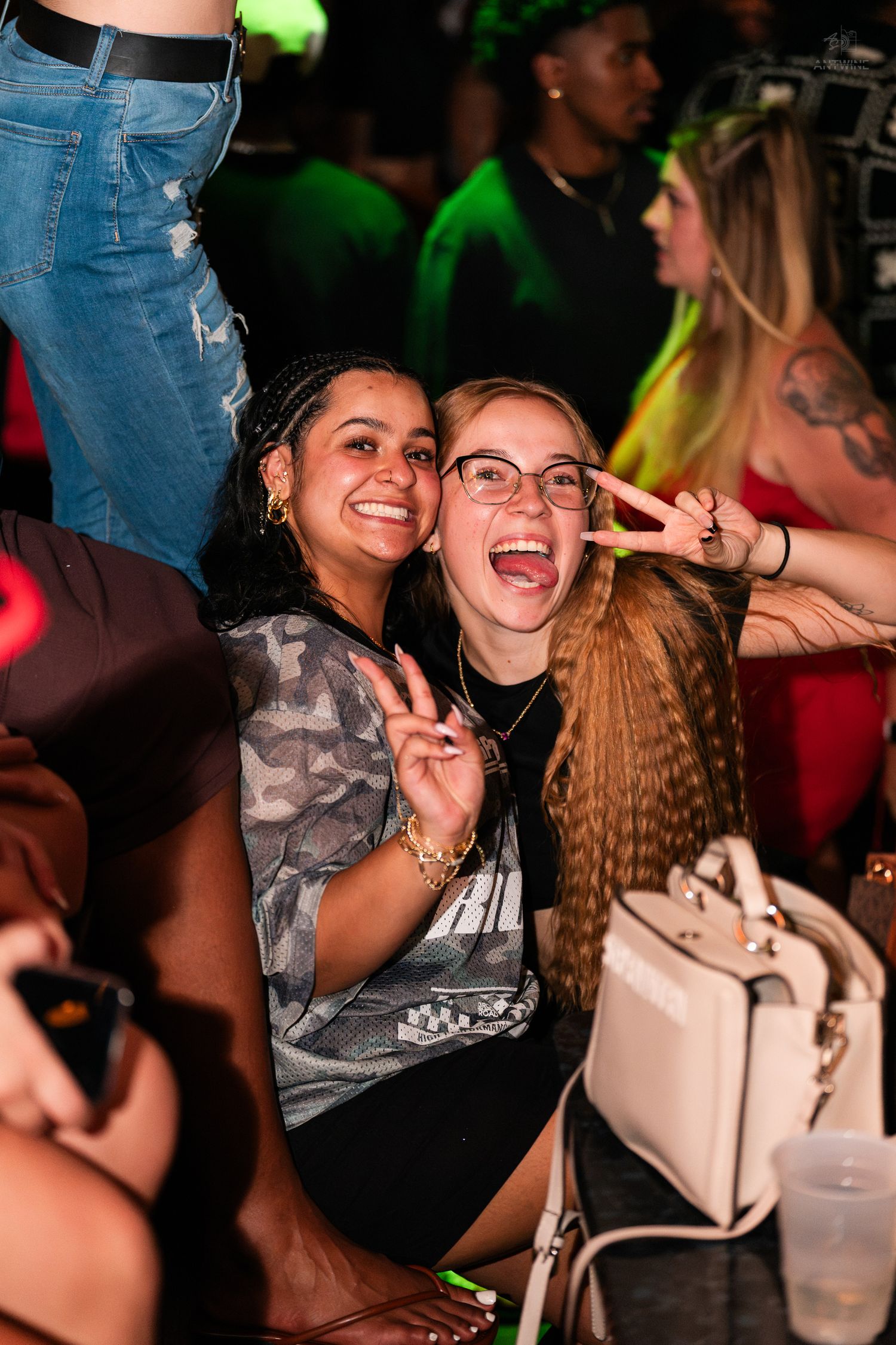 Two smiling women pose with peace signs at a nightclub, one wearing glasses holding a white purse.