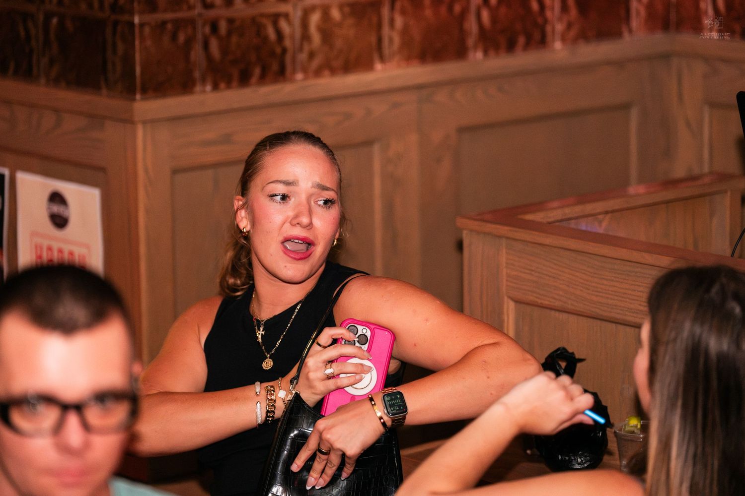 Young woman in black top holding a pink phone at a bar event.
