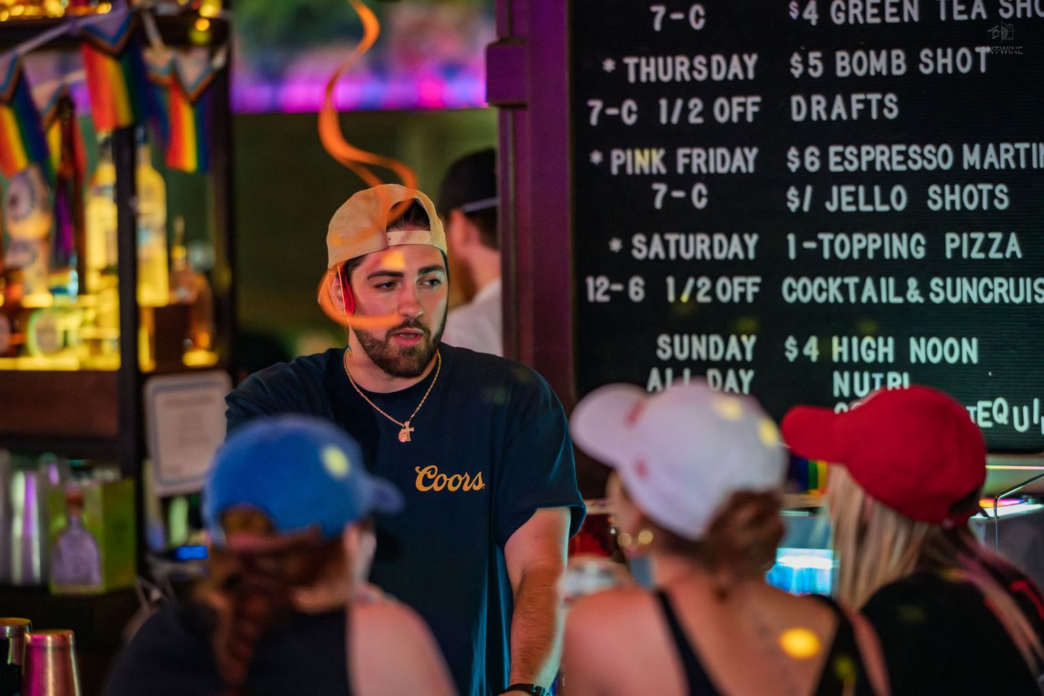 Bartender in Coors shirt stands at outdoor bar with colorful drink specials menu board.