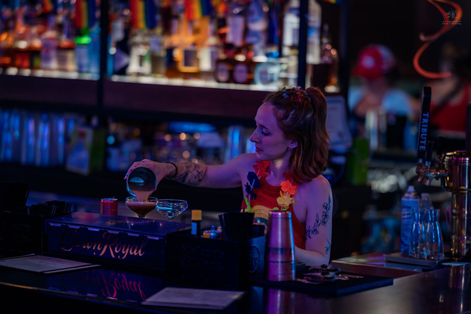 Tattooed female bartender pouring a drink at a neon-lit bar with Crown Royal branding.
