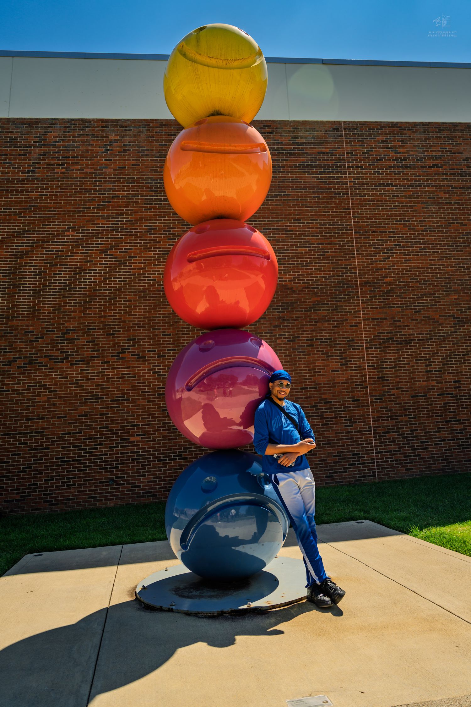 A person in a blue outfit leans against a large blue sphere beneath a tall rainbow-colored ball sculpture.