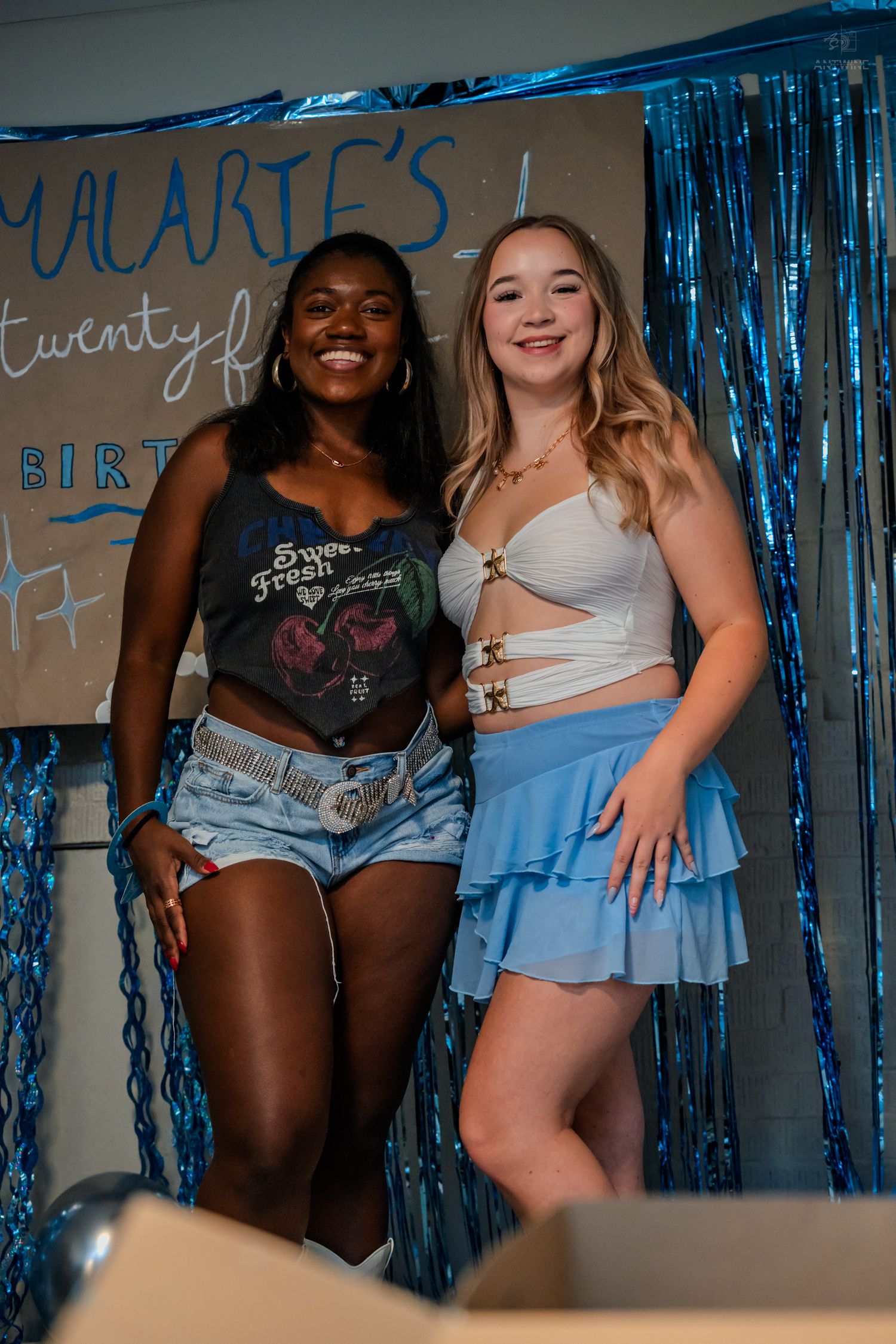 Two smiling women pose at Malarie's 21st birthday party with blue tinsel curtain backdrop.