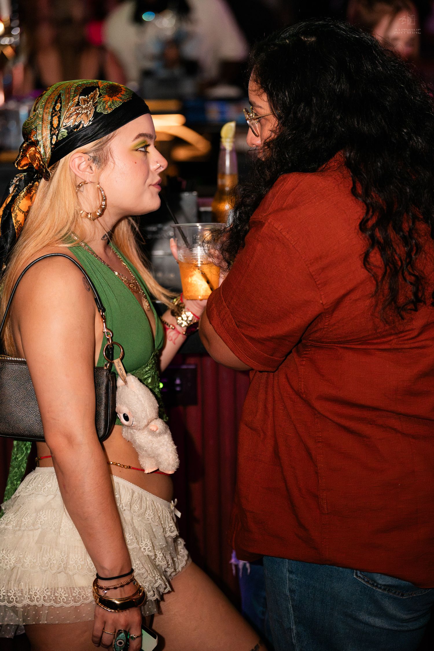A blonde woman in boho costume holds a stuffed animal while chatting with a woman in red at a bar.