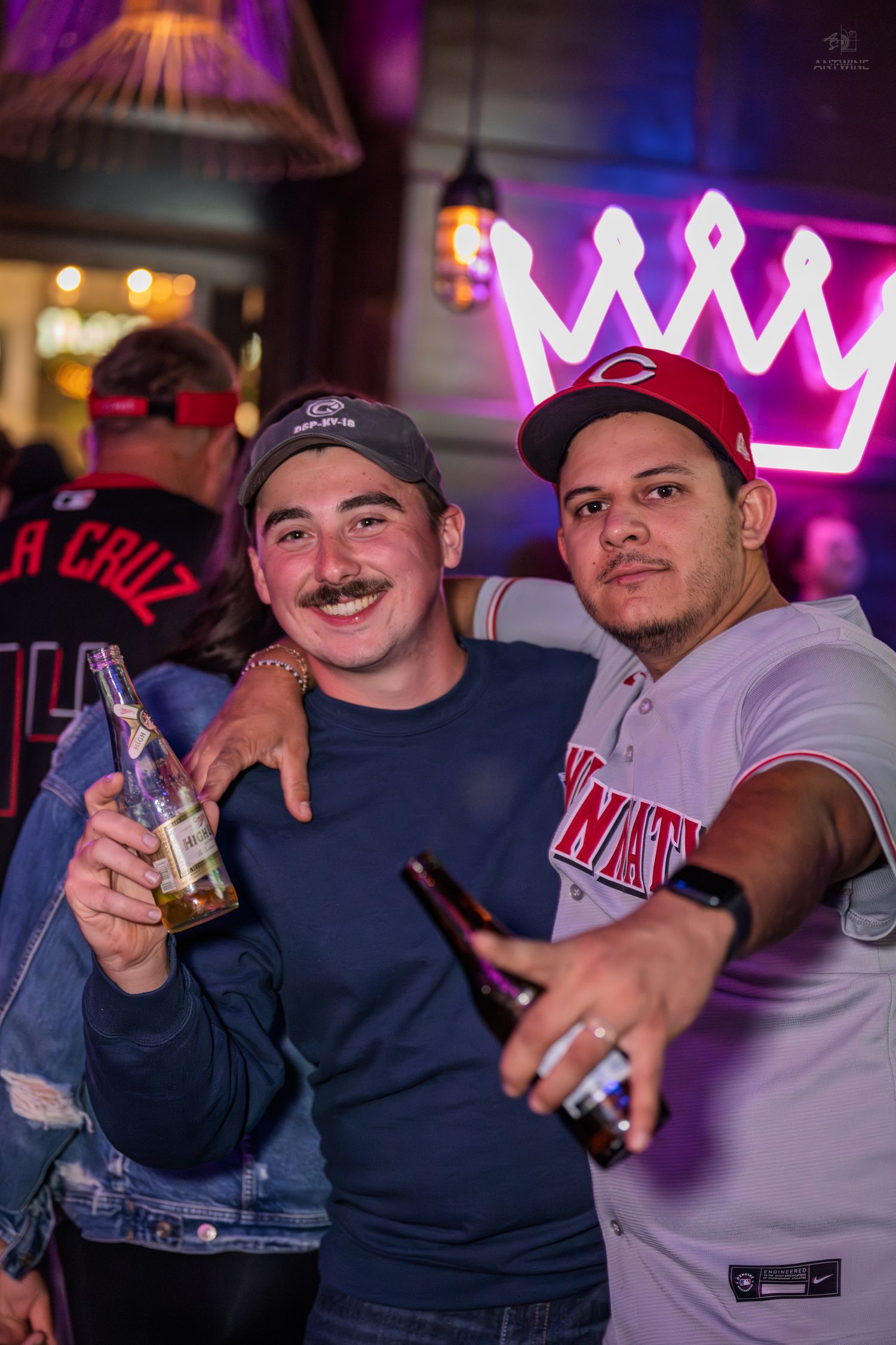 Two men smiling and holding beer bottles under a neon crown sign at a bar.