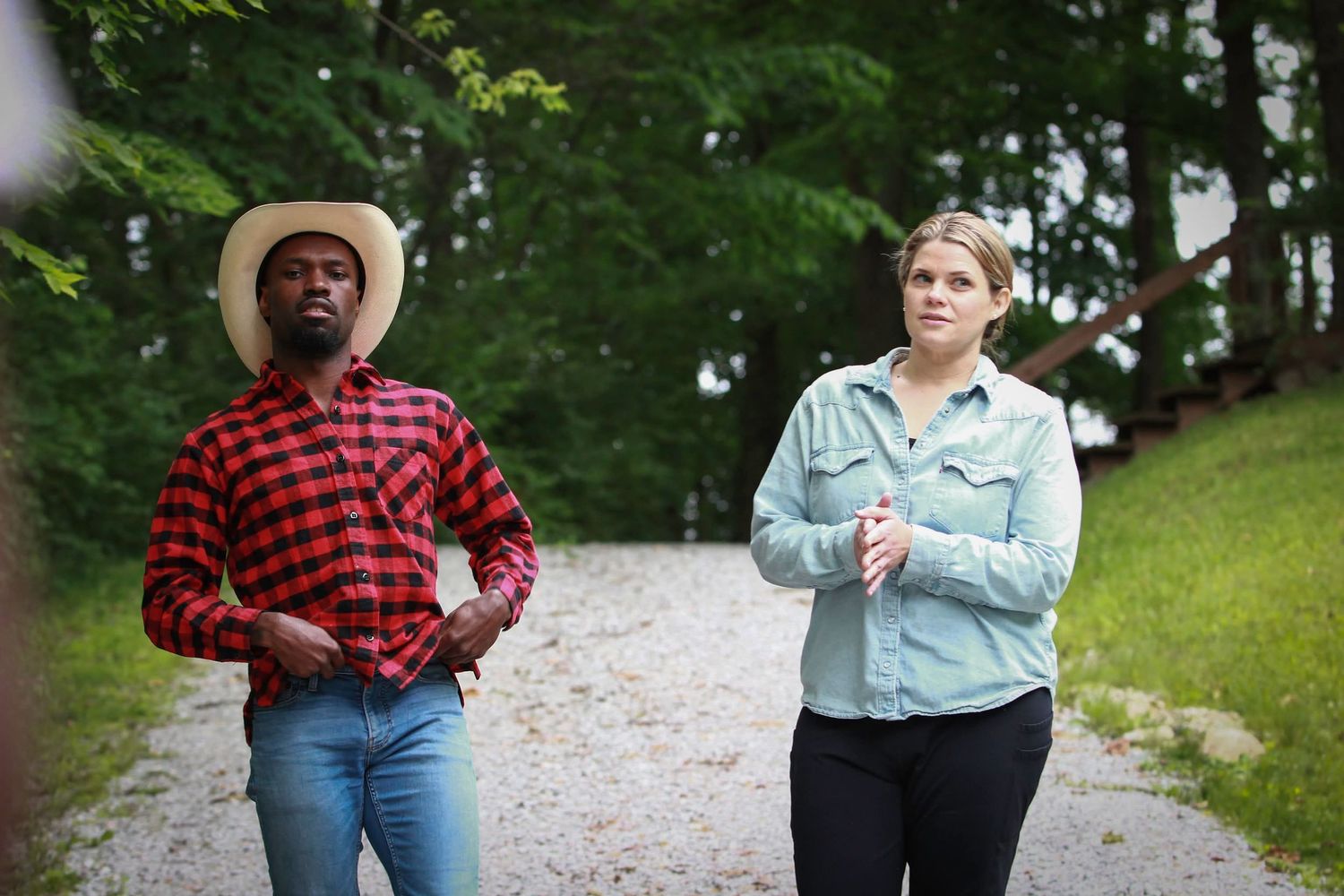 Man in red plaid shirt and straw hat walking with woman in denim shirt on a gravel path.