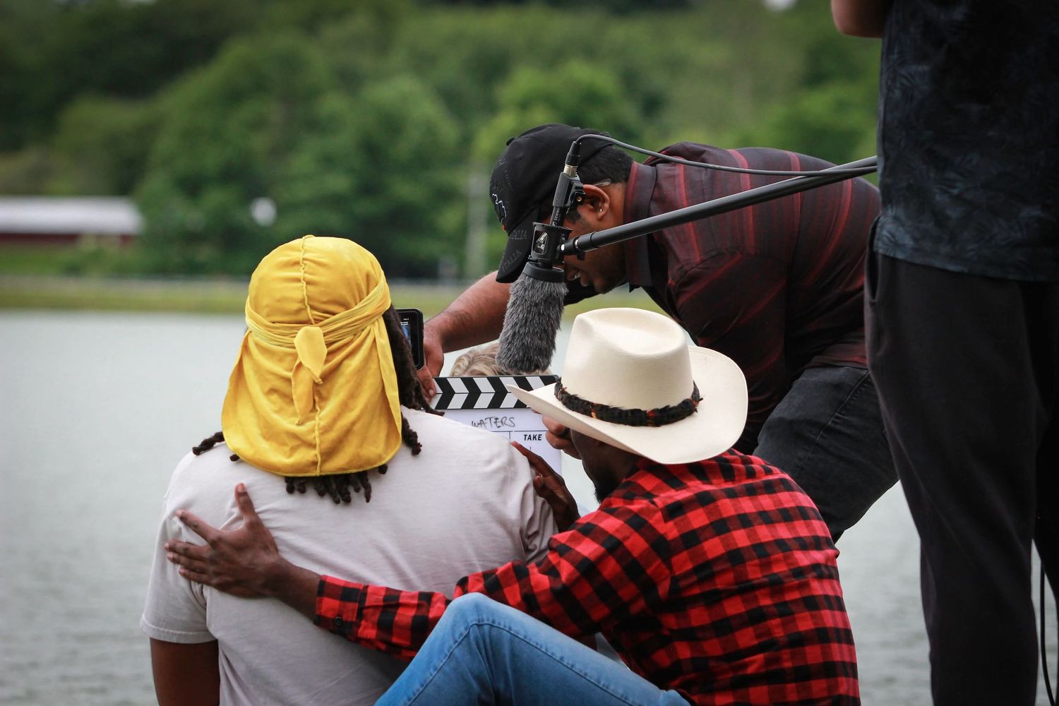 Three people wearing hats sit by a lake while a cameraman films them outdoors.