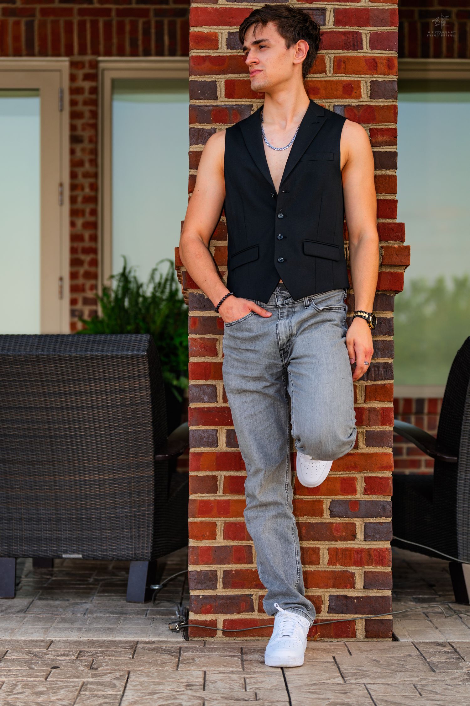 Young man in black vest and gray jeans leaning against brick column on outdoor patio.