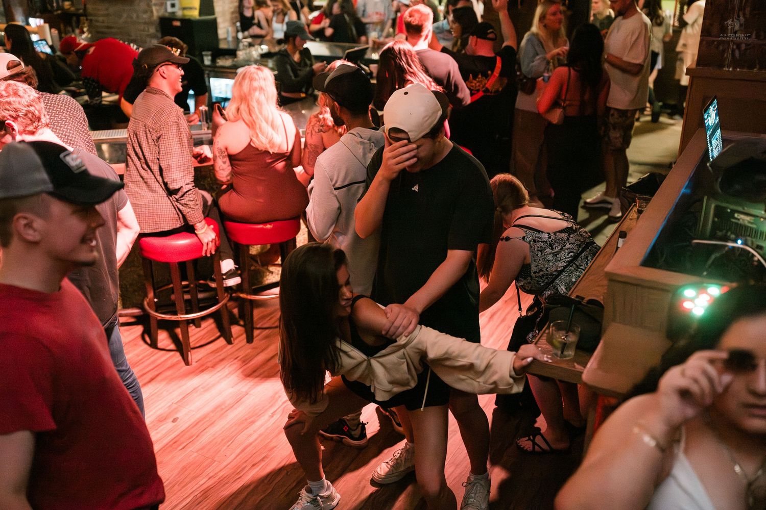 Lively bar crowd with people mingling and dancing under warm red lighting in a busy nightlife setting.