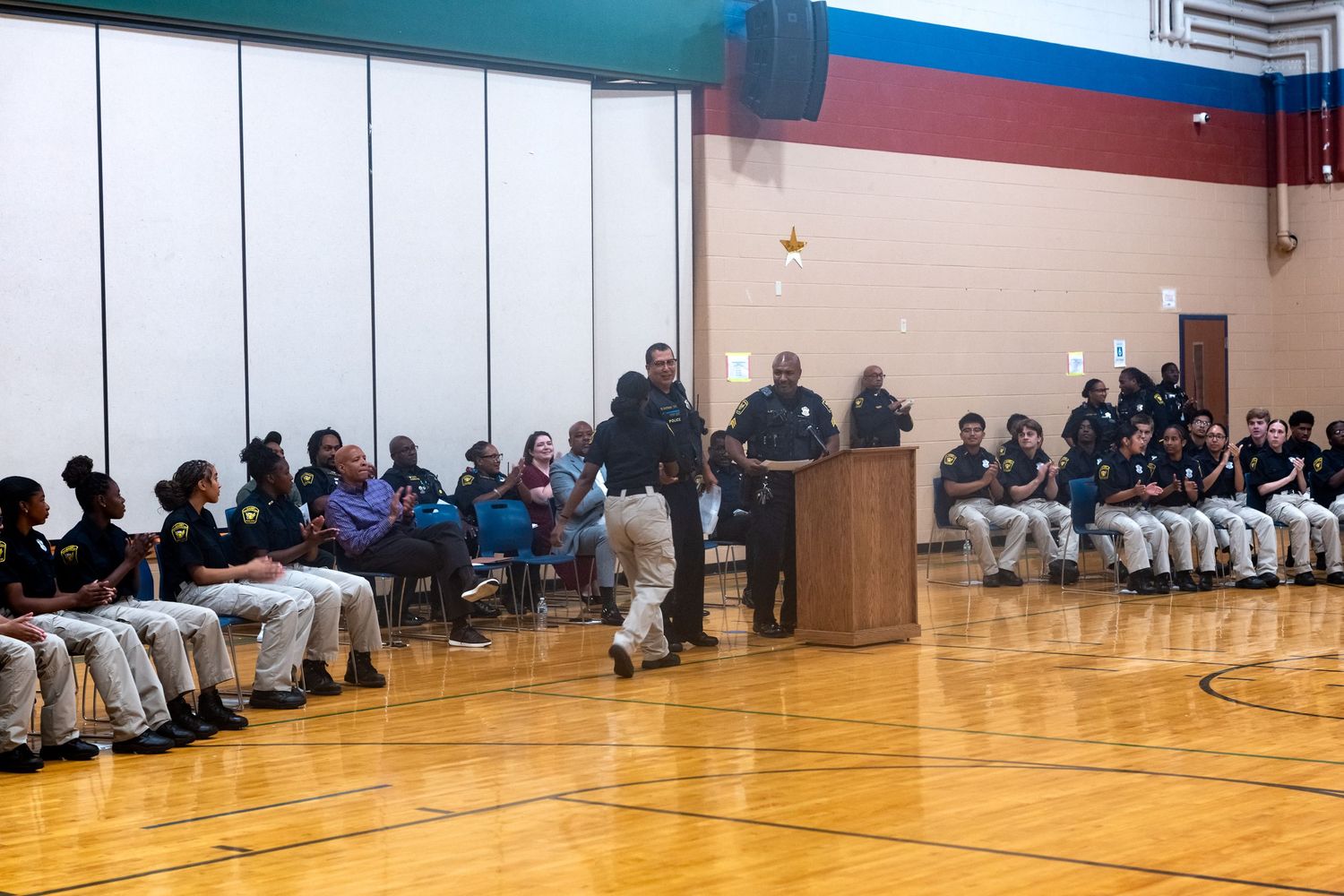 Students in uniforms sit in a school gymnasium during an assembly with speakers at a podium.