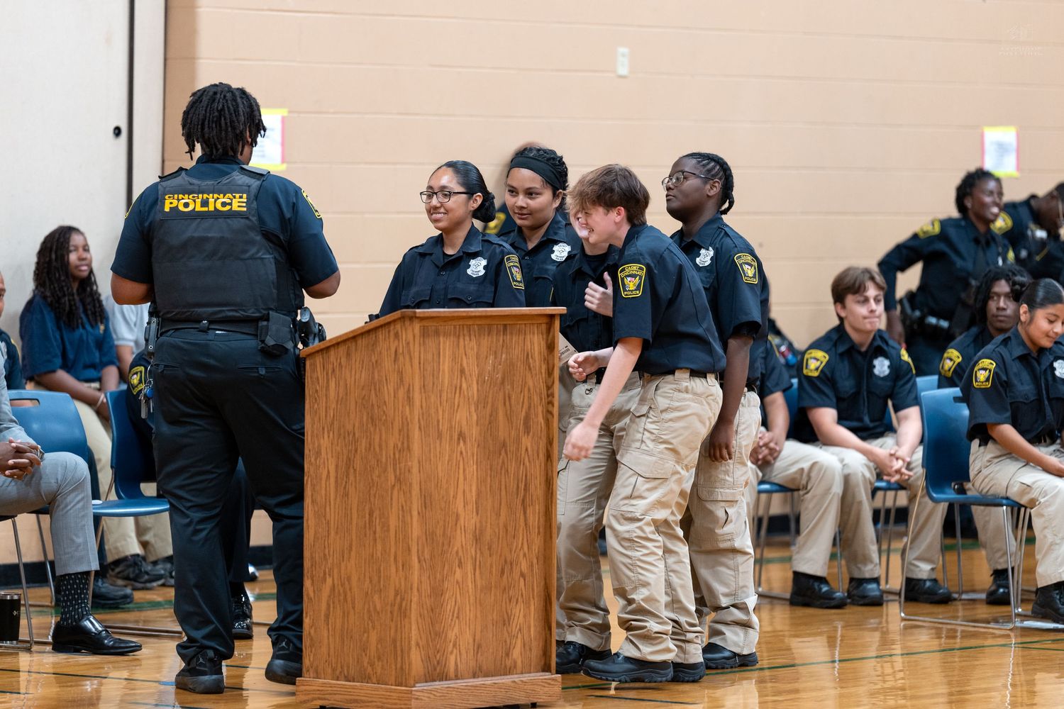 A police officer speaks with smiling youth cadets in uniform at an indoor school gymnasium event.