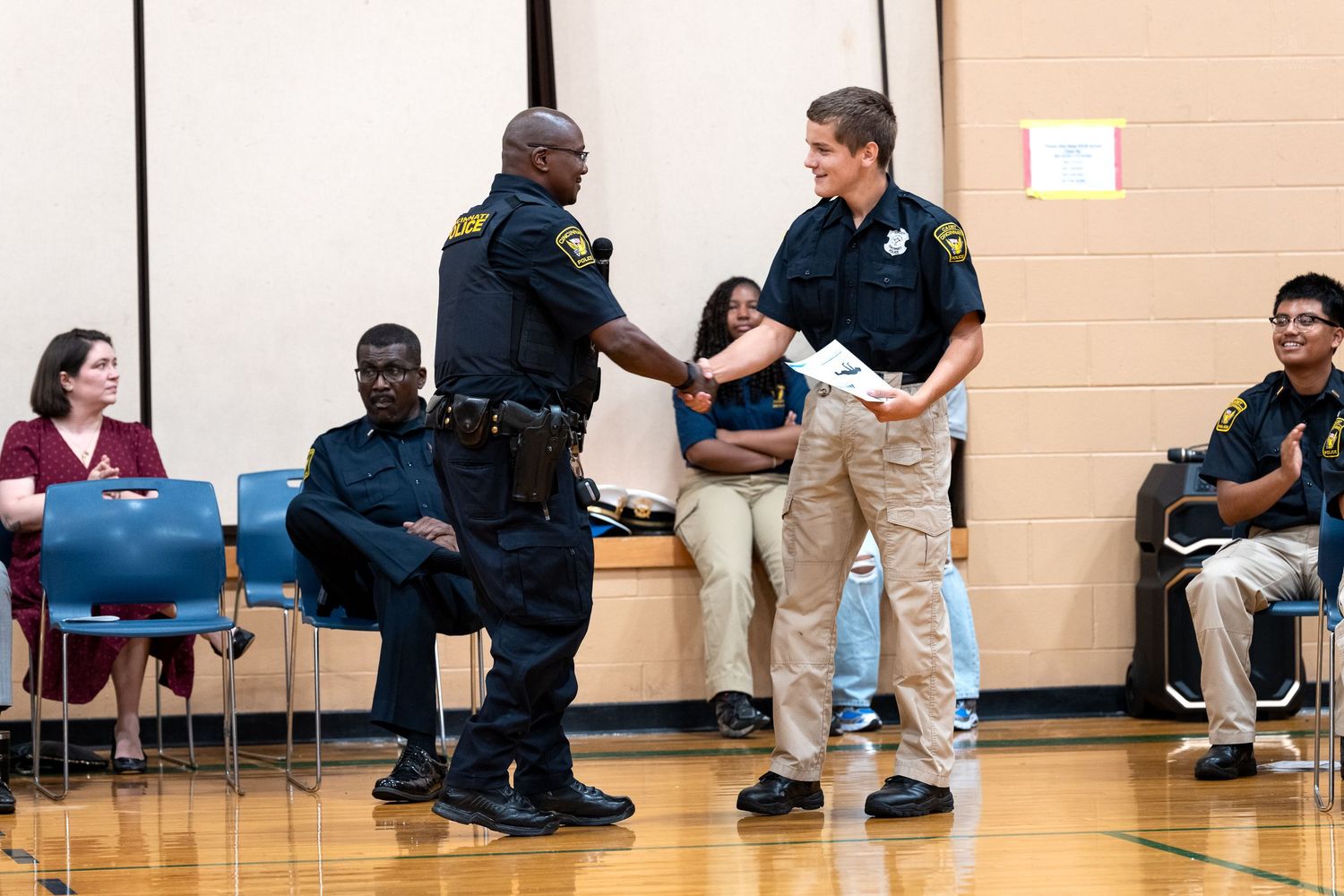 Two police officers shake hands during an awards ceremony inside a school gymnasium with attendees seated nearby.