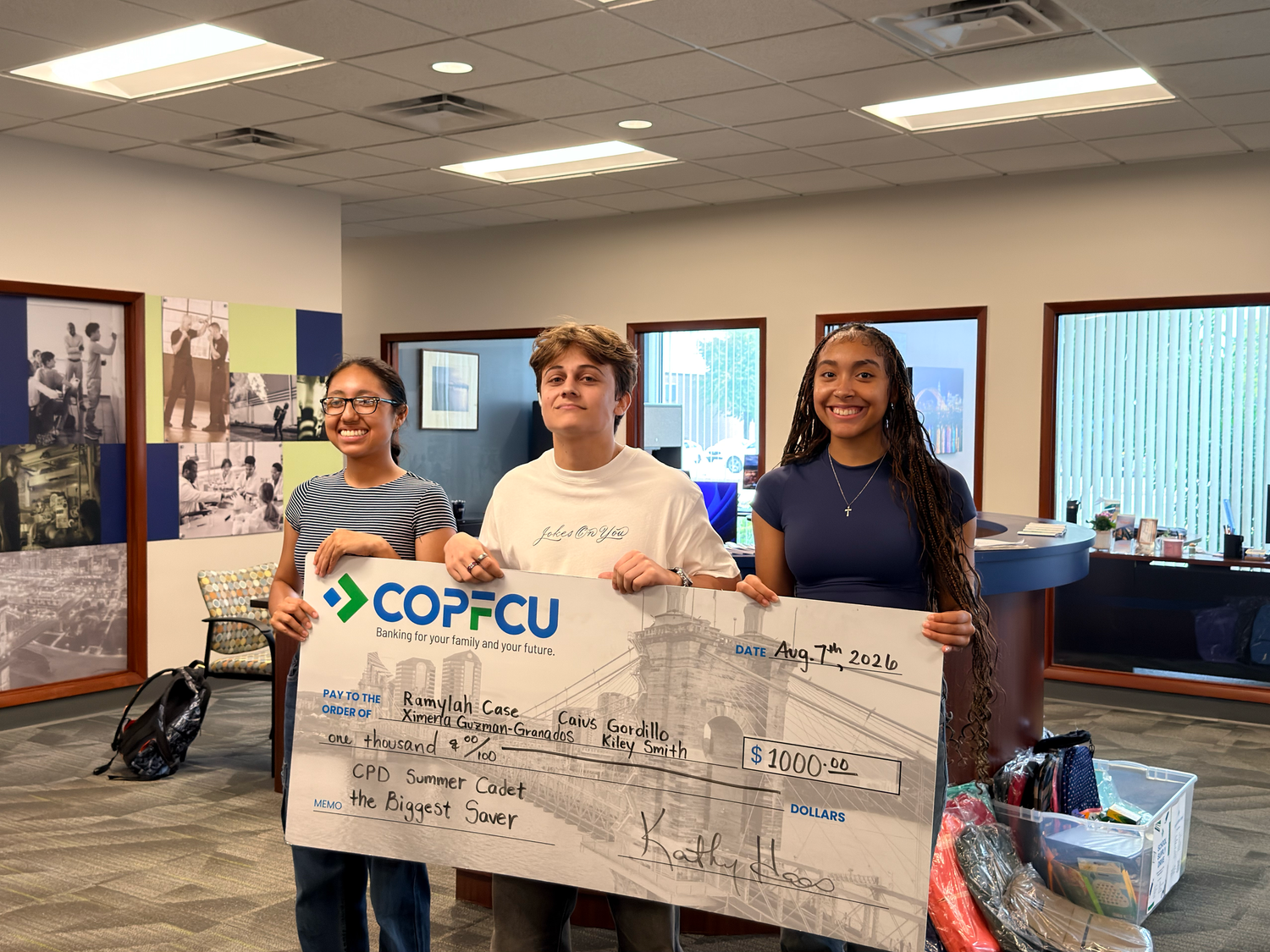 Three students hold a large COPFCU ceremonial check for $1,000 at a credit union branch office.