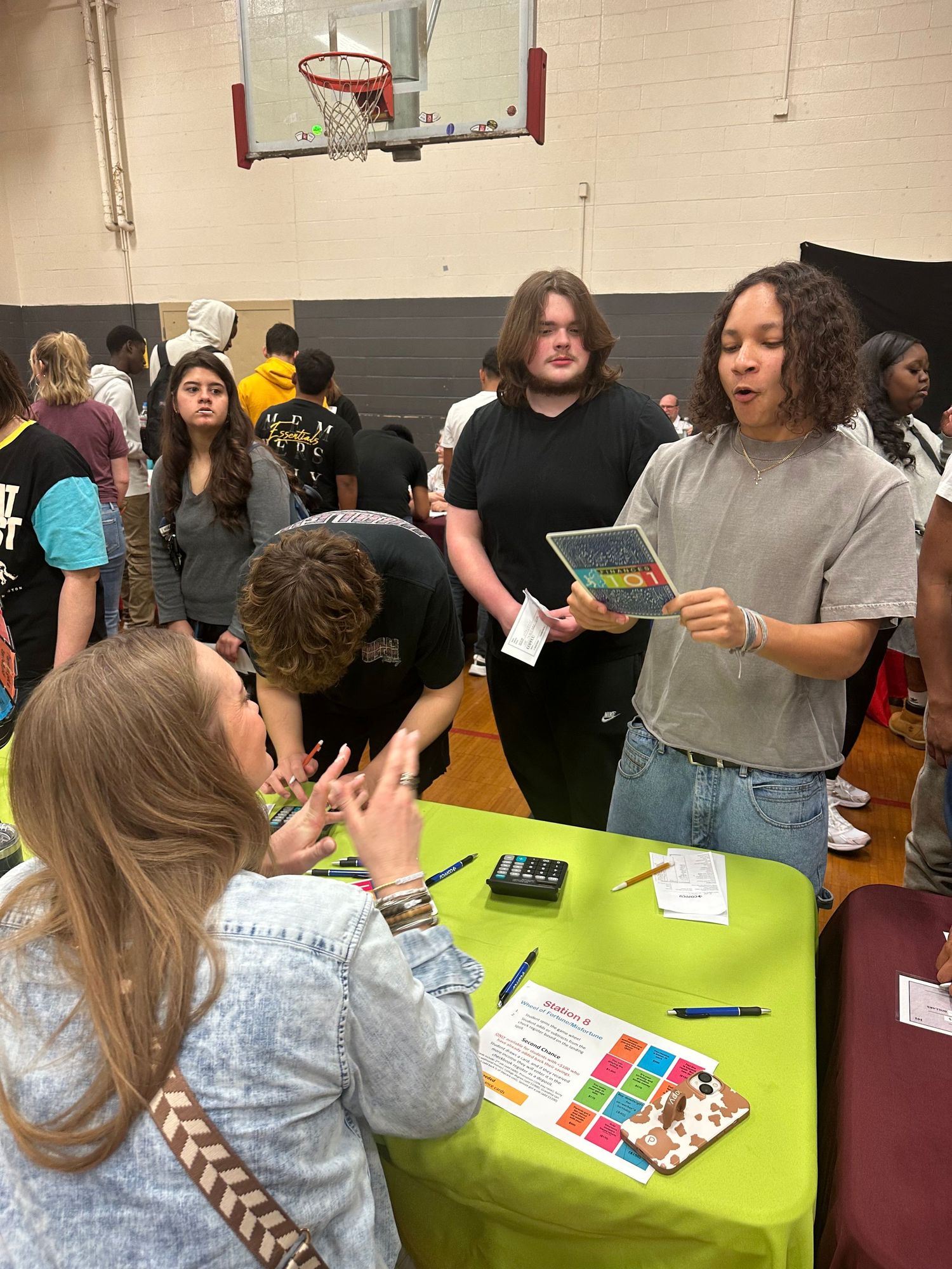 Students gather around a bright green table at a school gymnasium fair, reviewing colorful brochures and materials.
