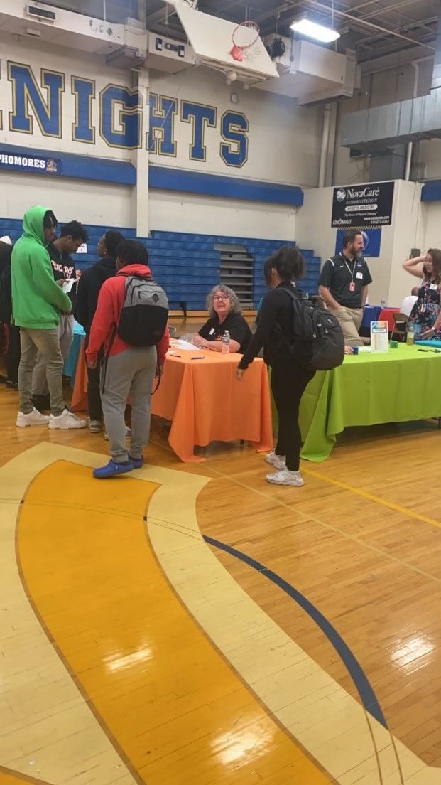 Students visit informational booths at a Knights school gymnasium career fair with colorful tables on a hardwood floor.