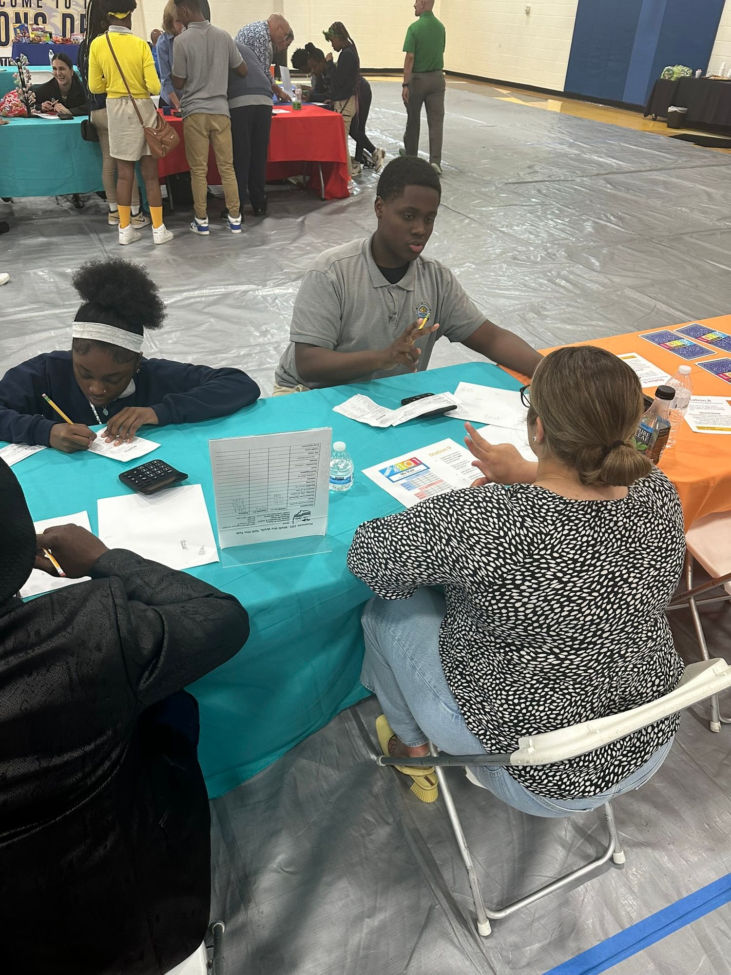 Students and adults sit at teal tables during a school career fair event held in a gymnasium.