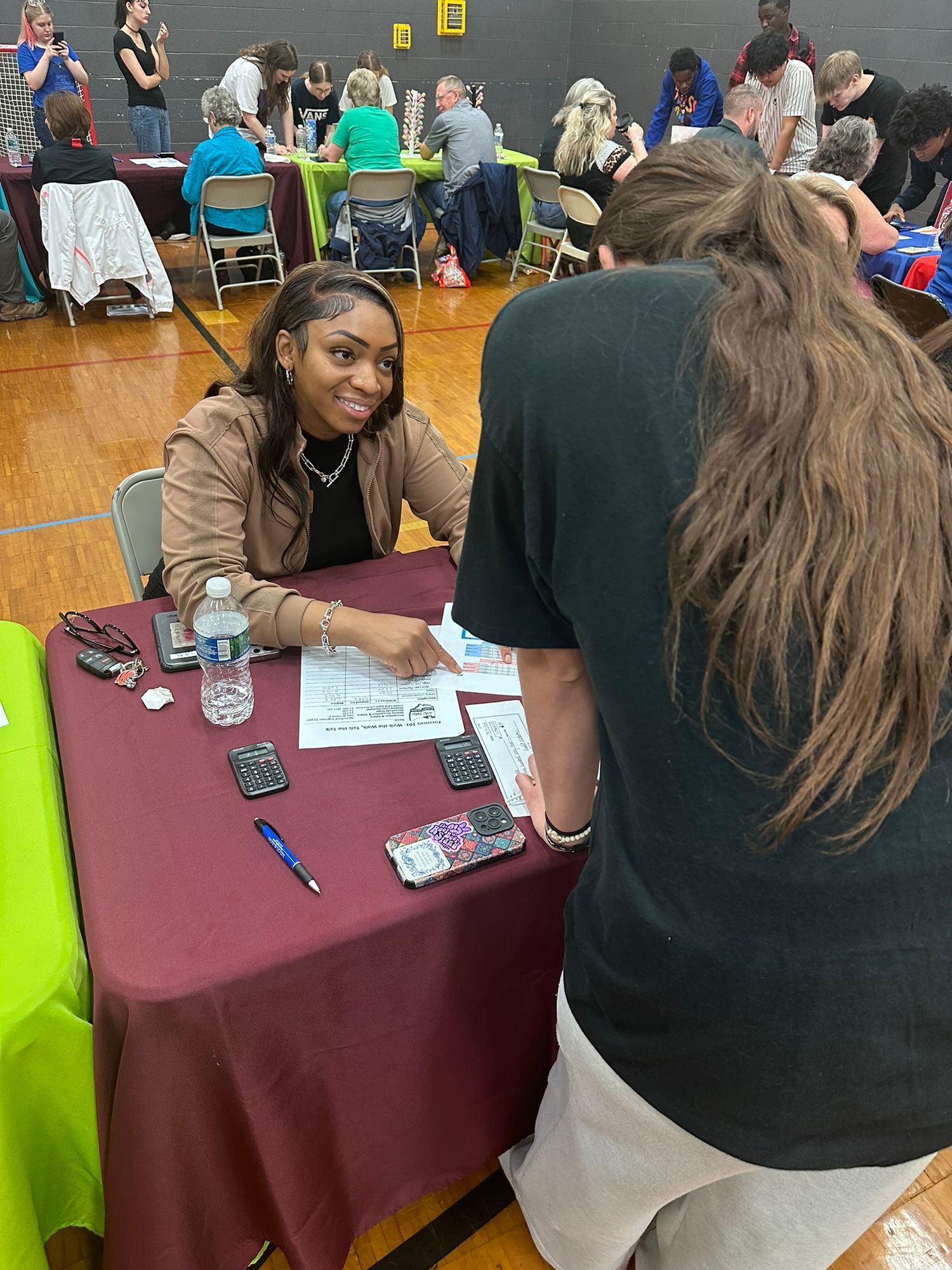 A woman sits at a maroon-draped table consulting with a visitor at an indoor community event or job fair.