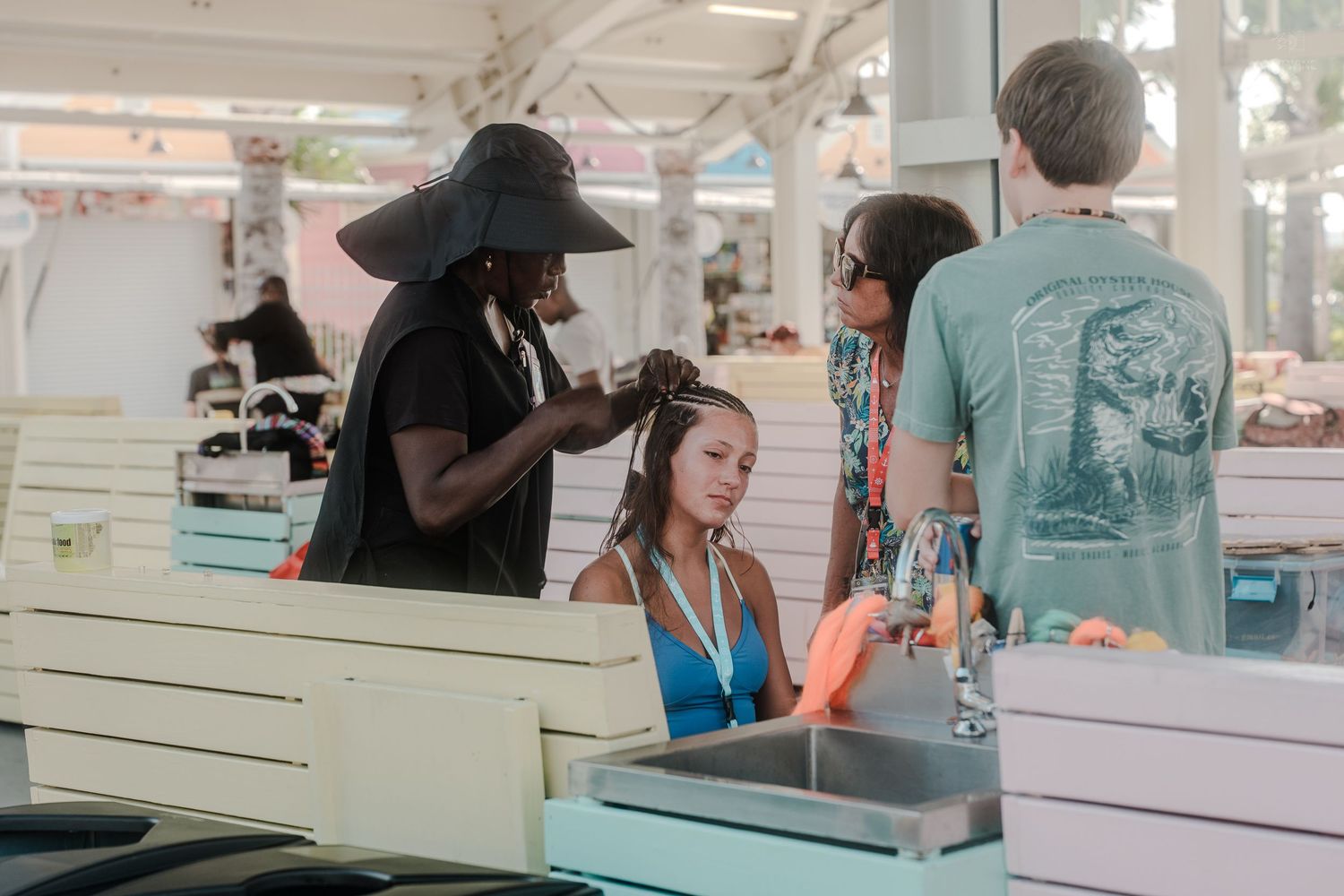 A hairstylist braiding a young girl's hair at an outdoor beach market stall on a sunny day.