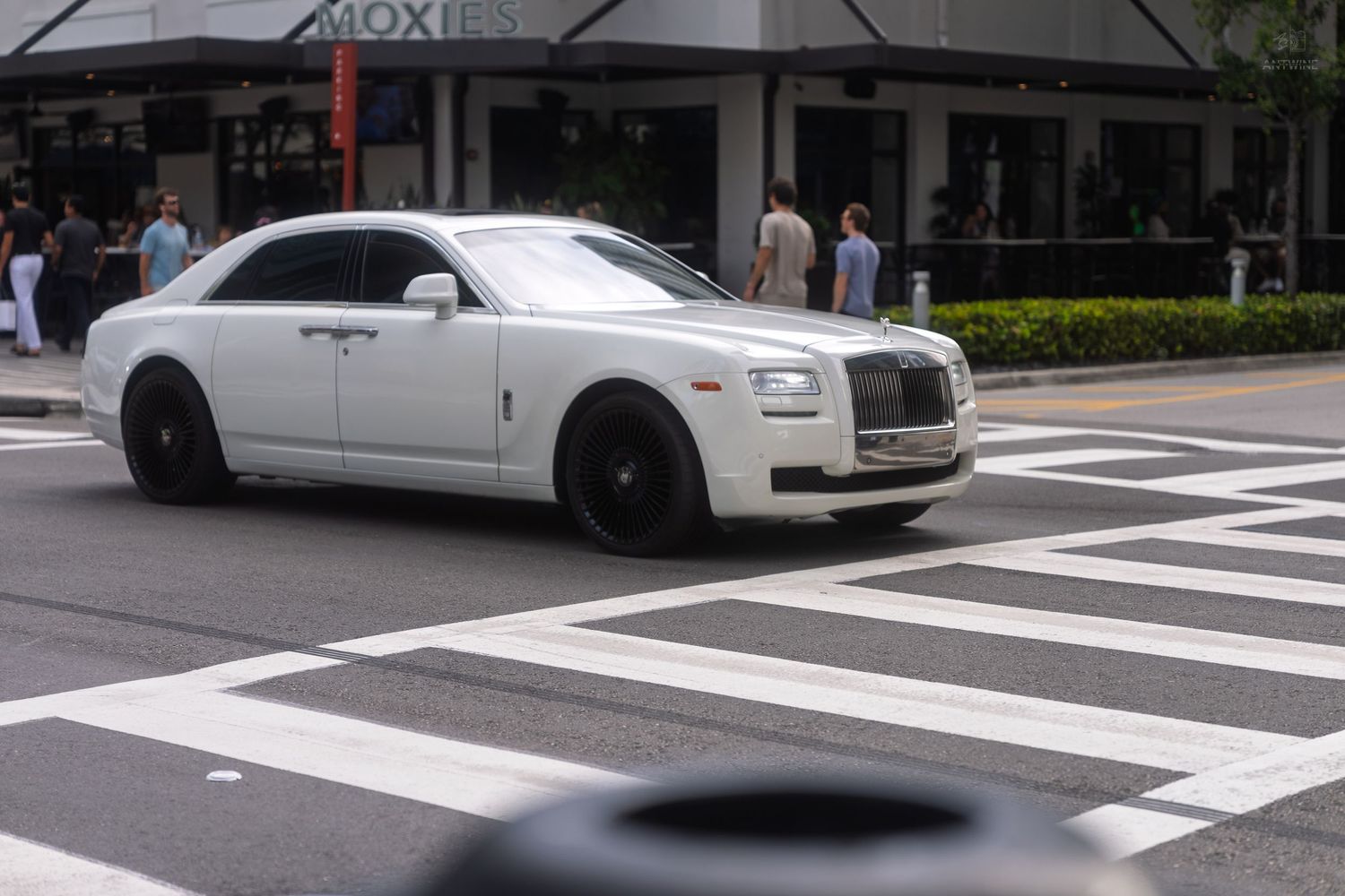 White Rolls-Royce Ghost luxury sedan crossing a striped pedestrian crosswalk on a sunny urban street.