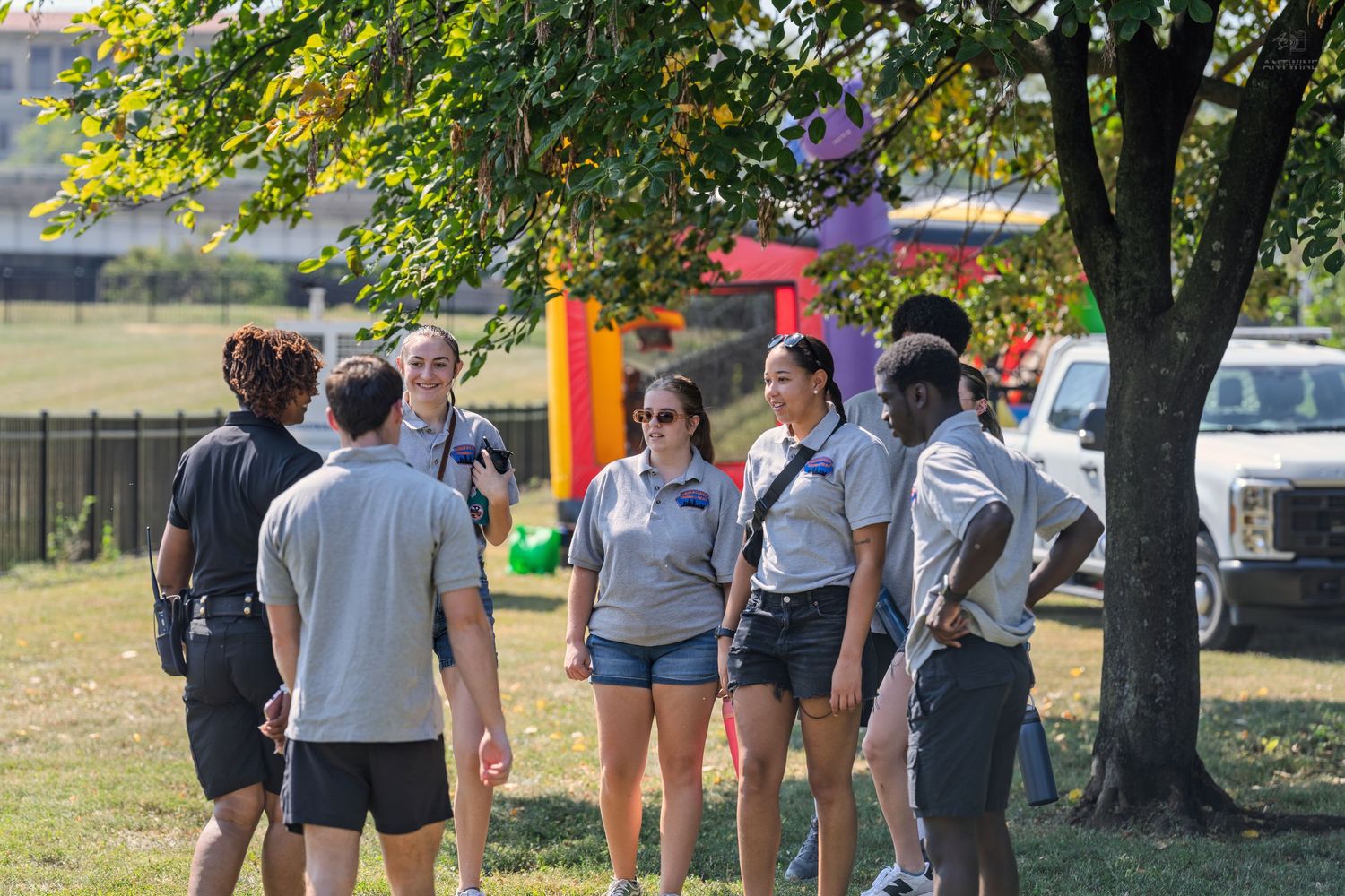Group of young adults in matching gray shirts gather outdoors near a tree at a colorful community event on a sunny day.