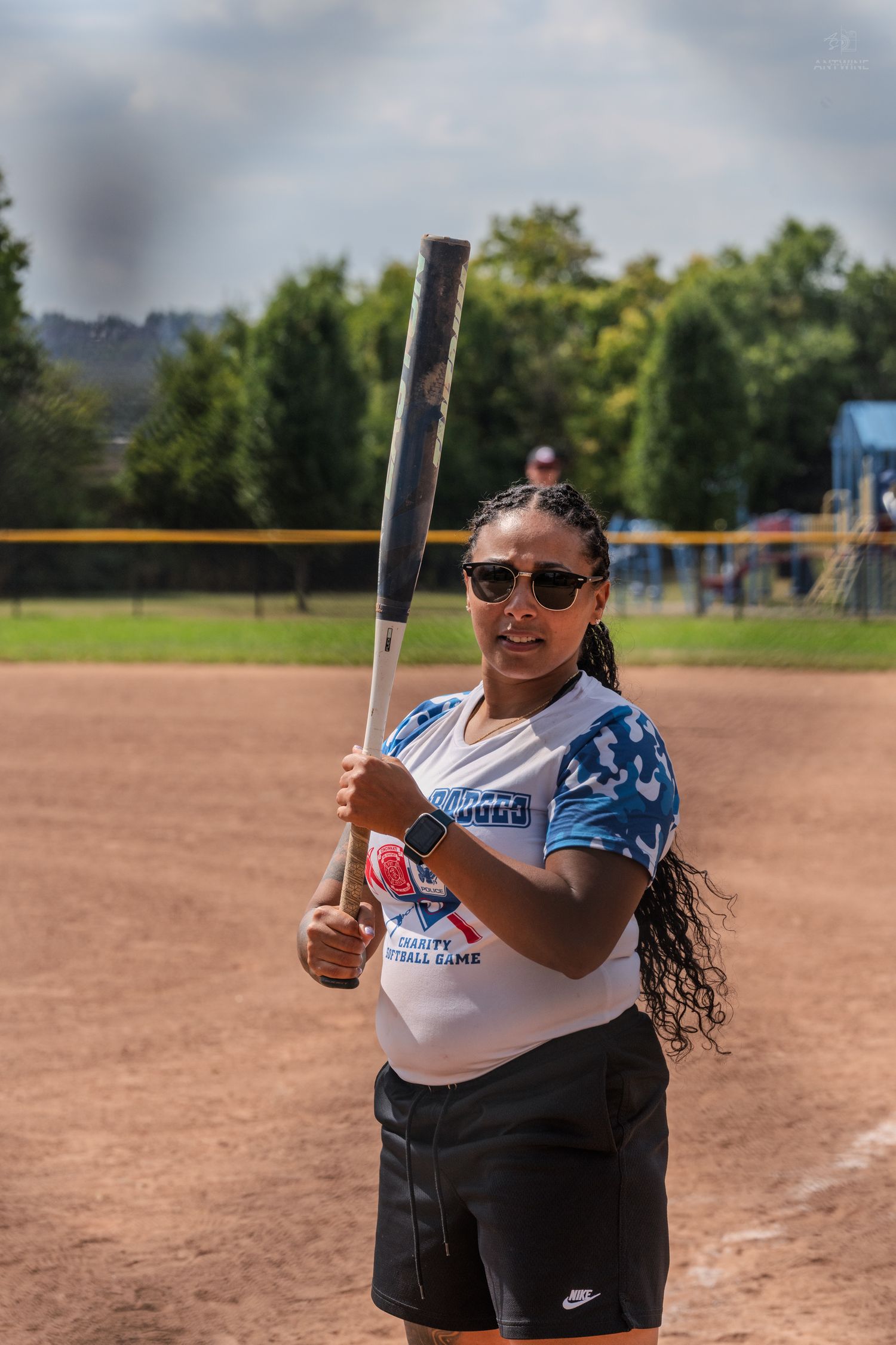 Woman in Battle of the Badges charity softball jersey holds a bat on a dirt field wearing sunglasses.