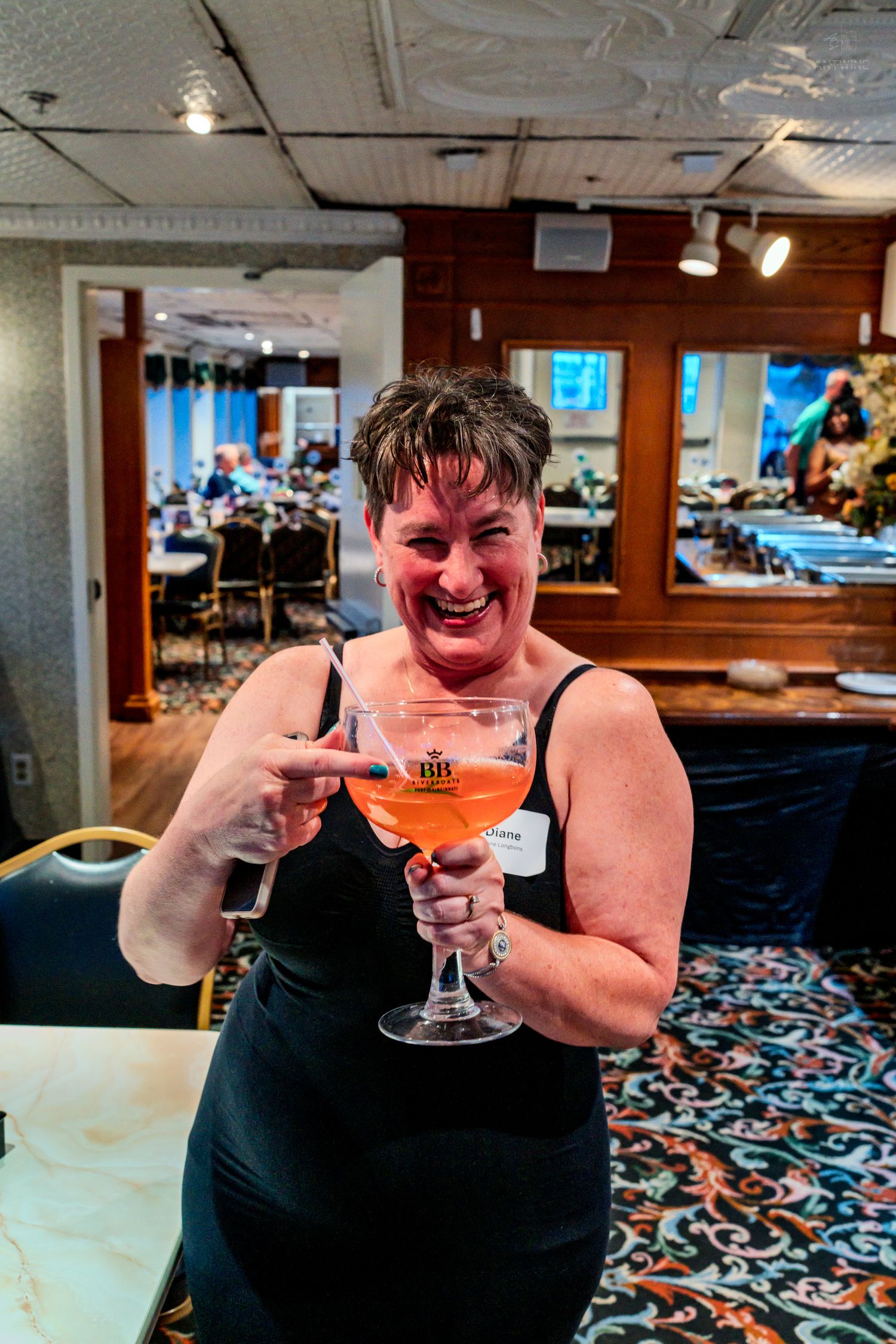 A laughing woman in a black dress holds two large wine glasses with orange cocktails at a restaurant event.