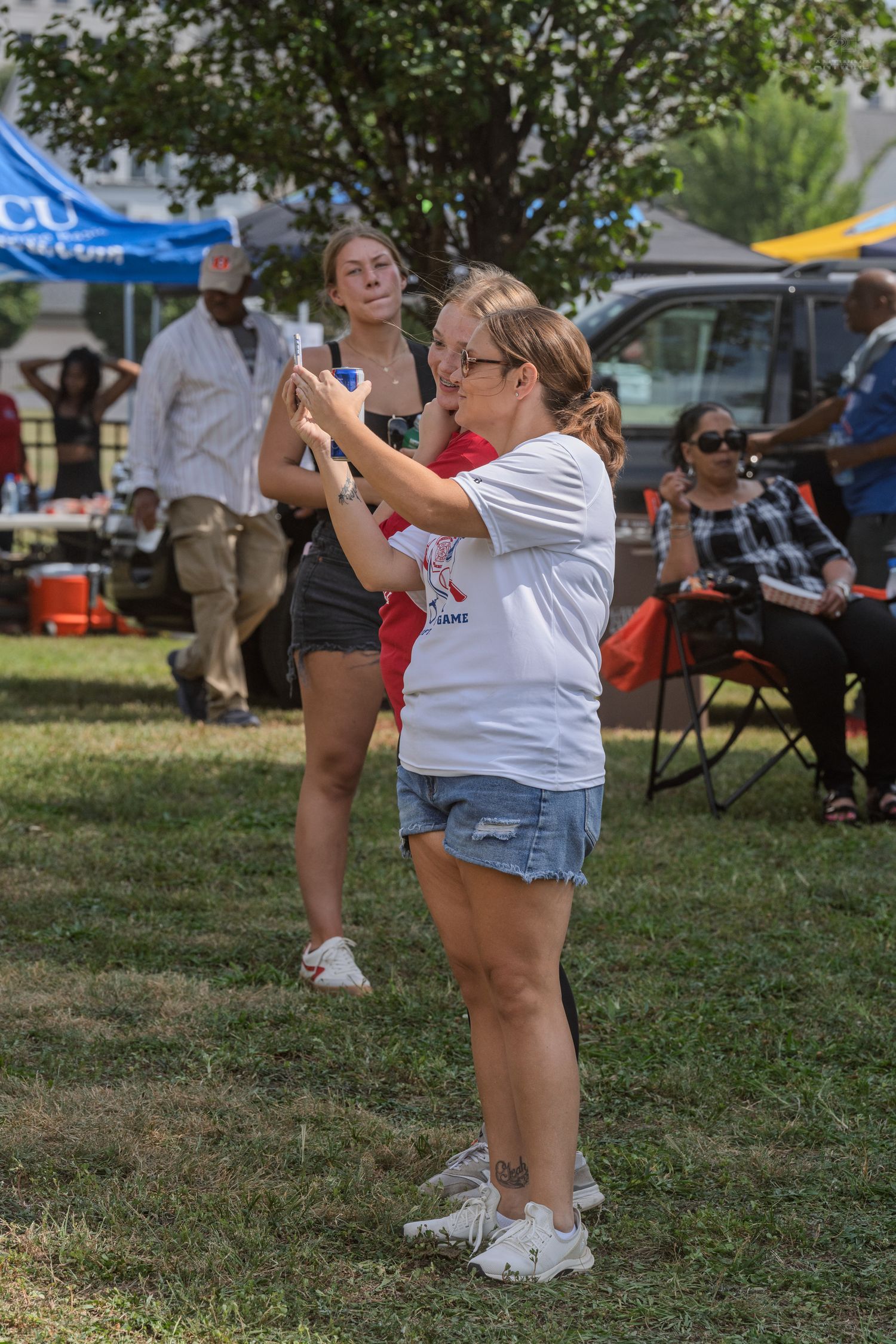 Woman in white t-shirt taking photos on smartphone at an outdoor summer community event.