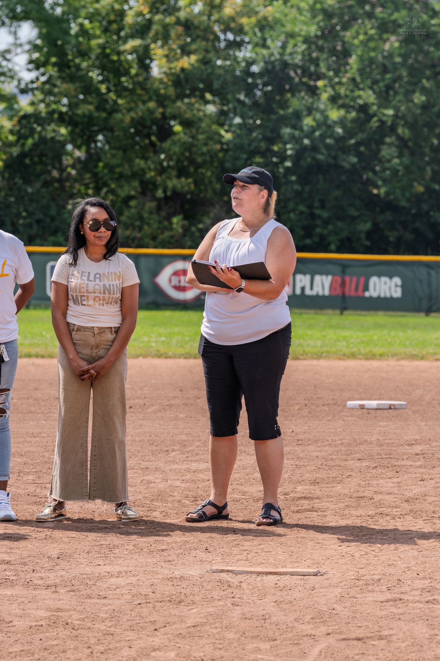 Two women stand on a baseball diamond during an outdoor ceremony on a sunny day.