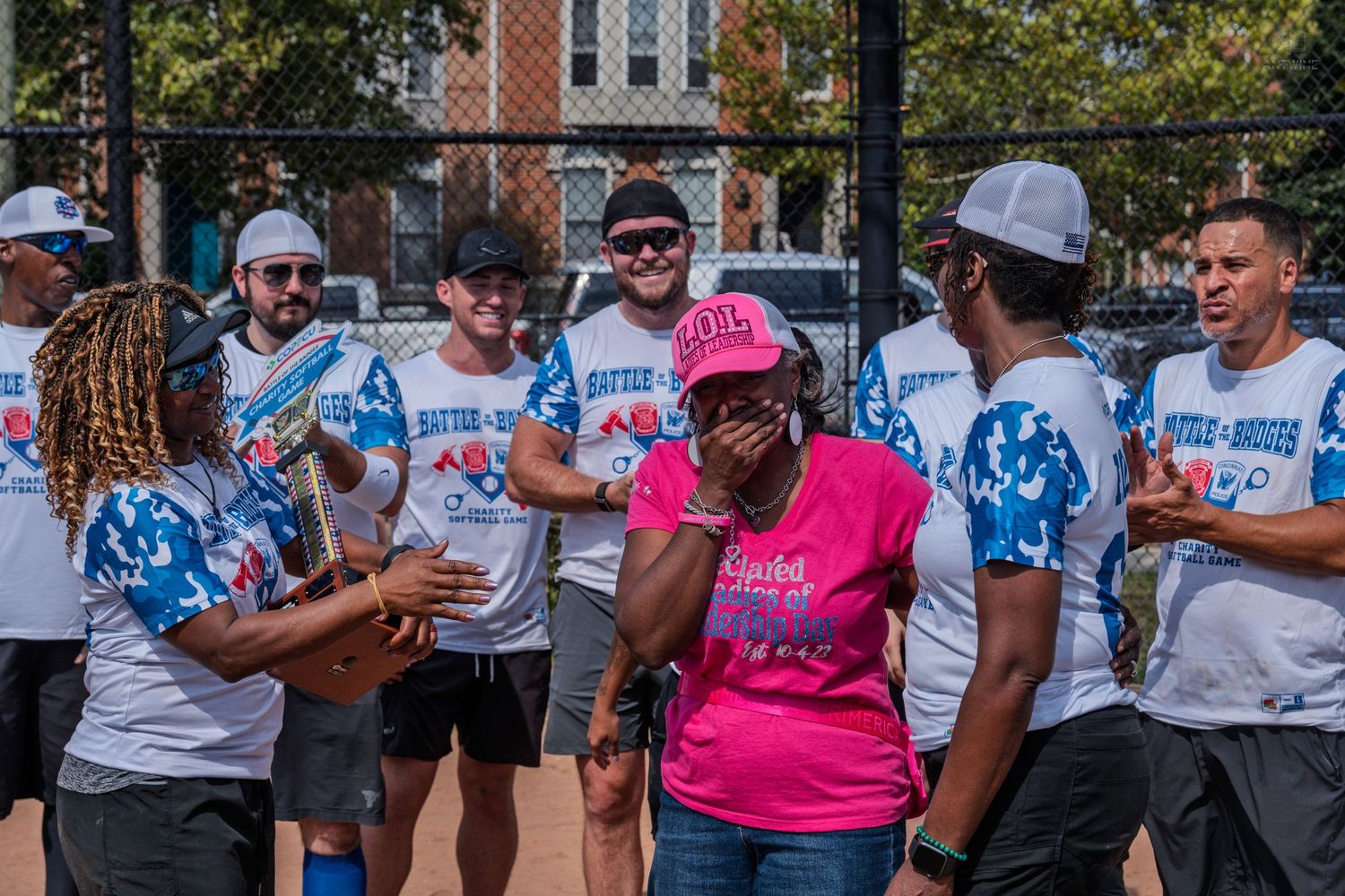 Emotional woman in pink shirt surrounded by smiling Battle of the Badges charity softball event participants outdoors.