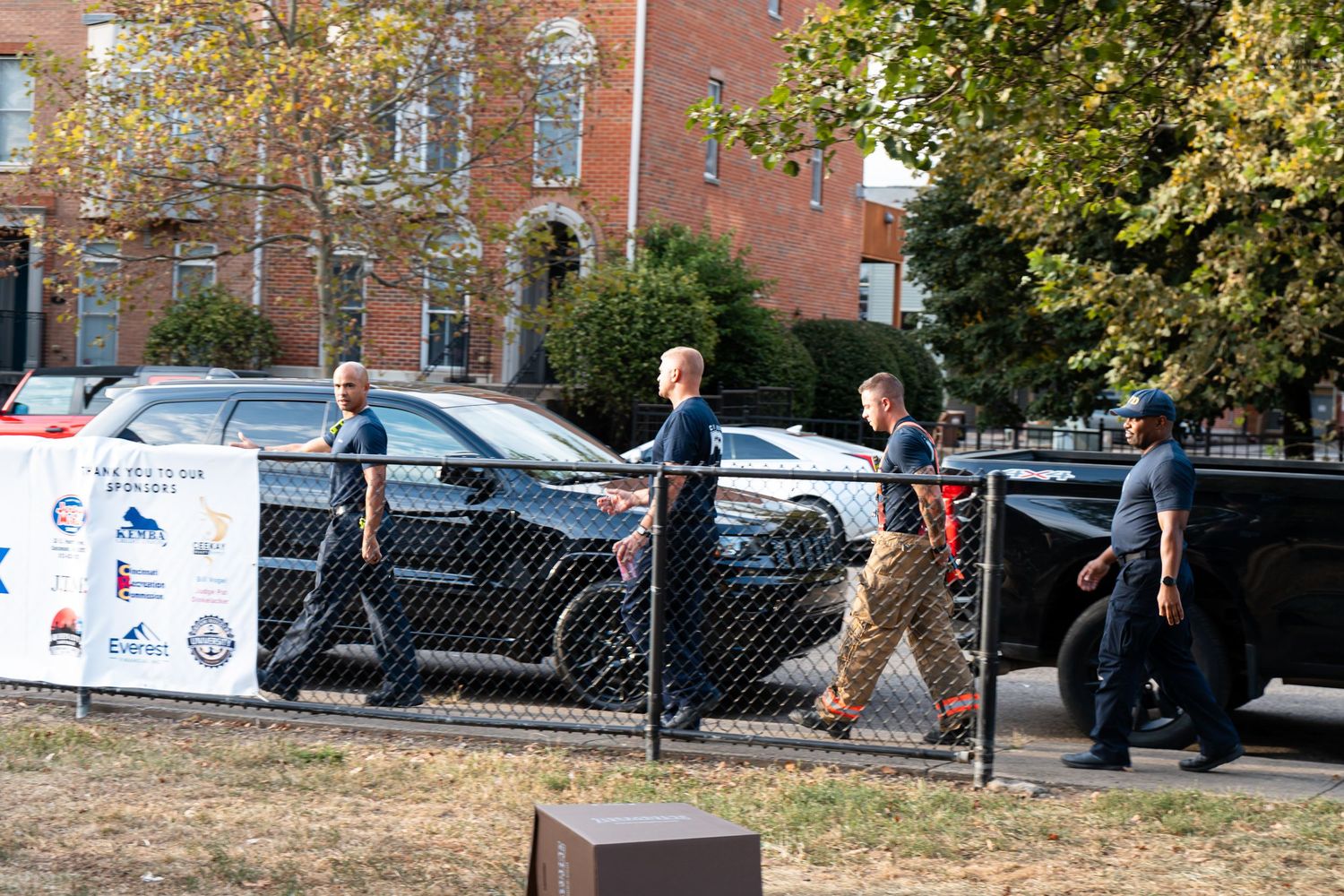 Group of people walking near parked cars behind a chain-link fence at an outdoor event.