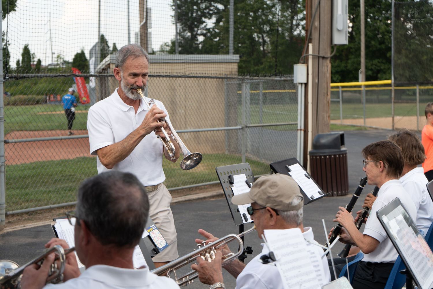 Musician plays trumpet while leading an outdoor band rehearsal at a baseball field.