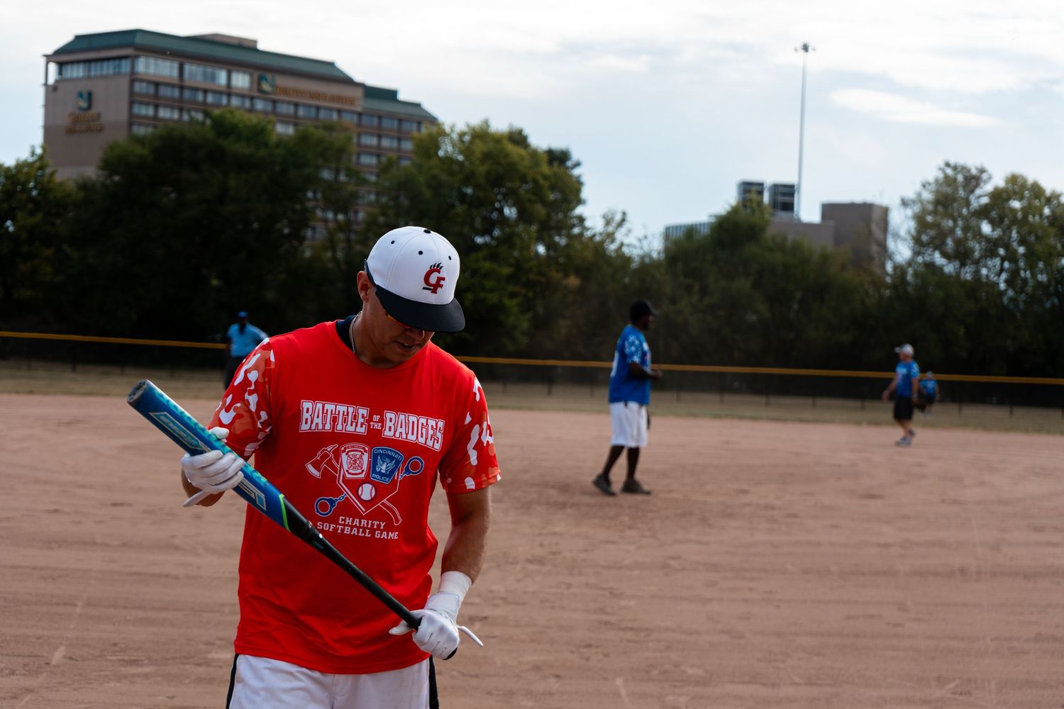 Softball player in red Battle of the Badges jersey holds a bat on the dirt field during a charity game.