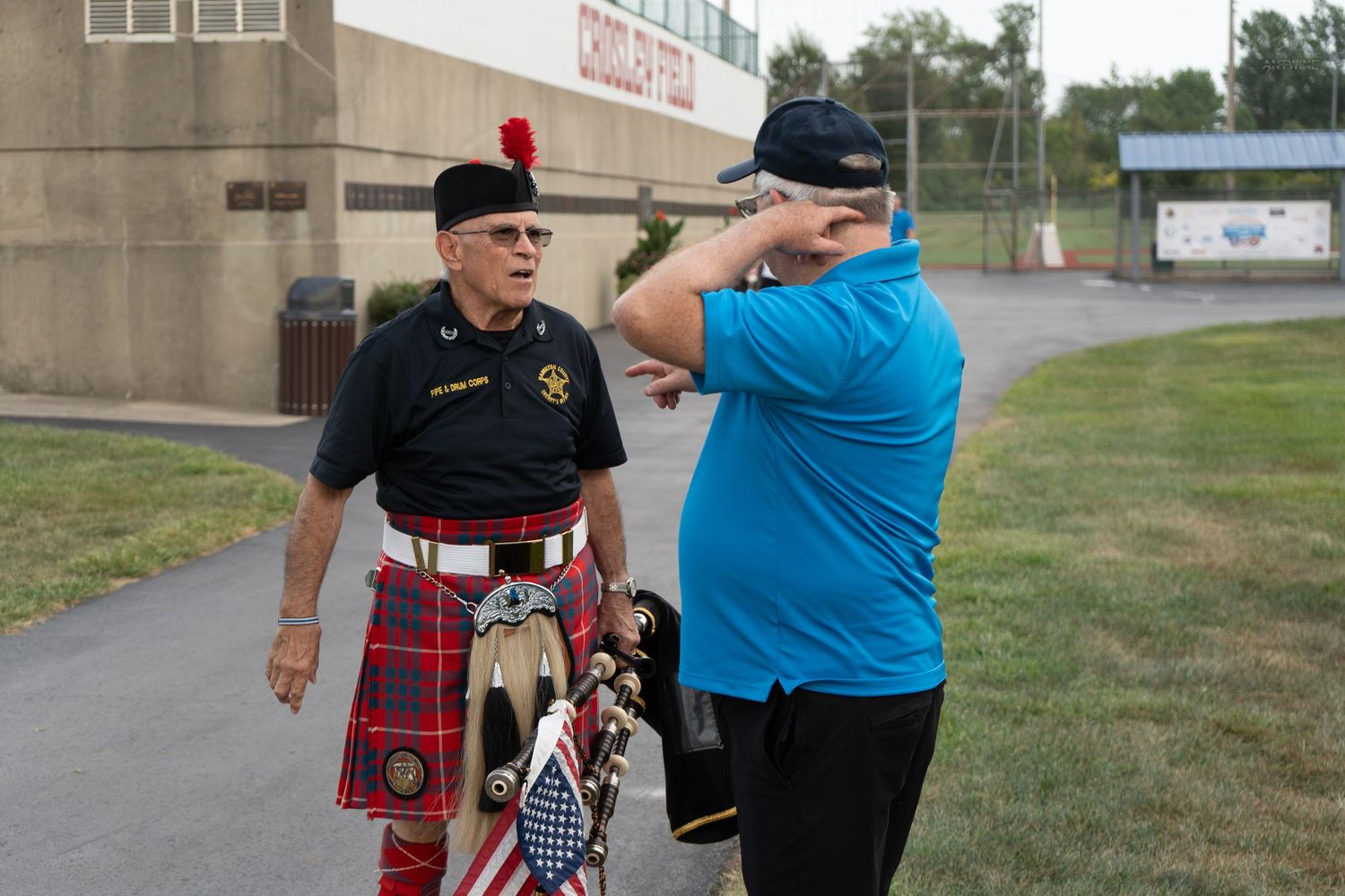 A man in a red kilt with bagpipes stands next to a person in a blue shirt at an outdoor event.