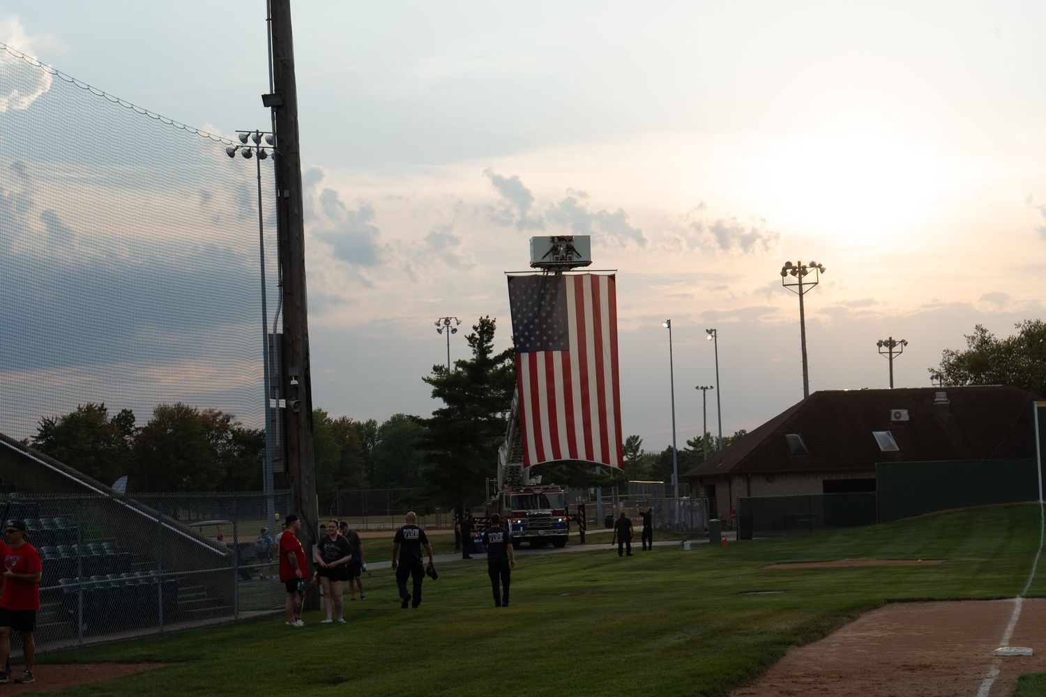 Baseball field at dusk with large American flag displayed on building in background under dramatic sunset sky.