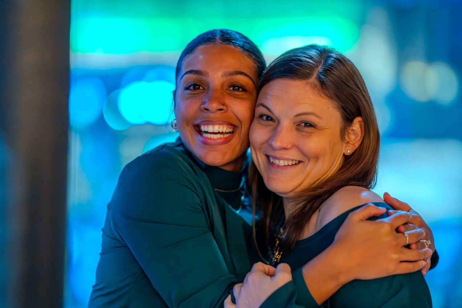 Two smiling women share a warm hug against a vibrant blue bokeh background.