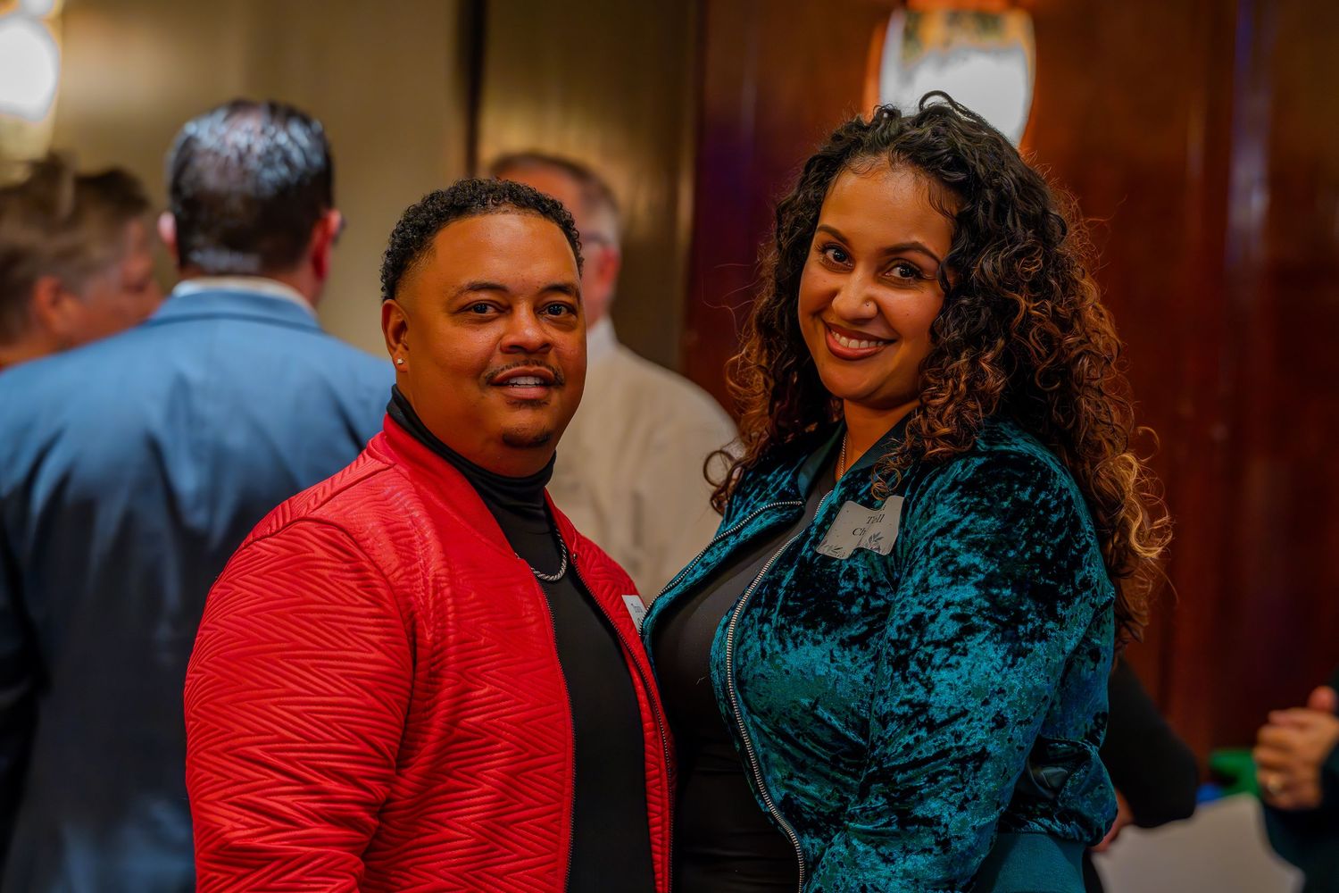 A man in a red jacket and a woman in a teal blazer smile together at an indoor social event.