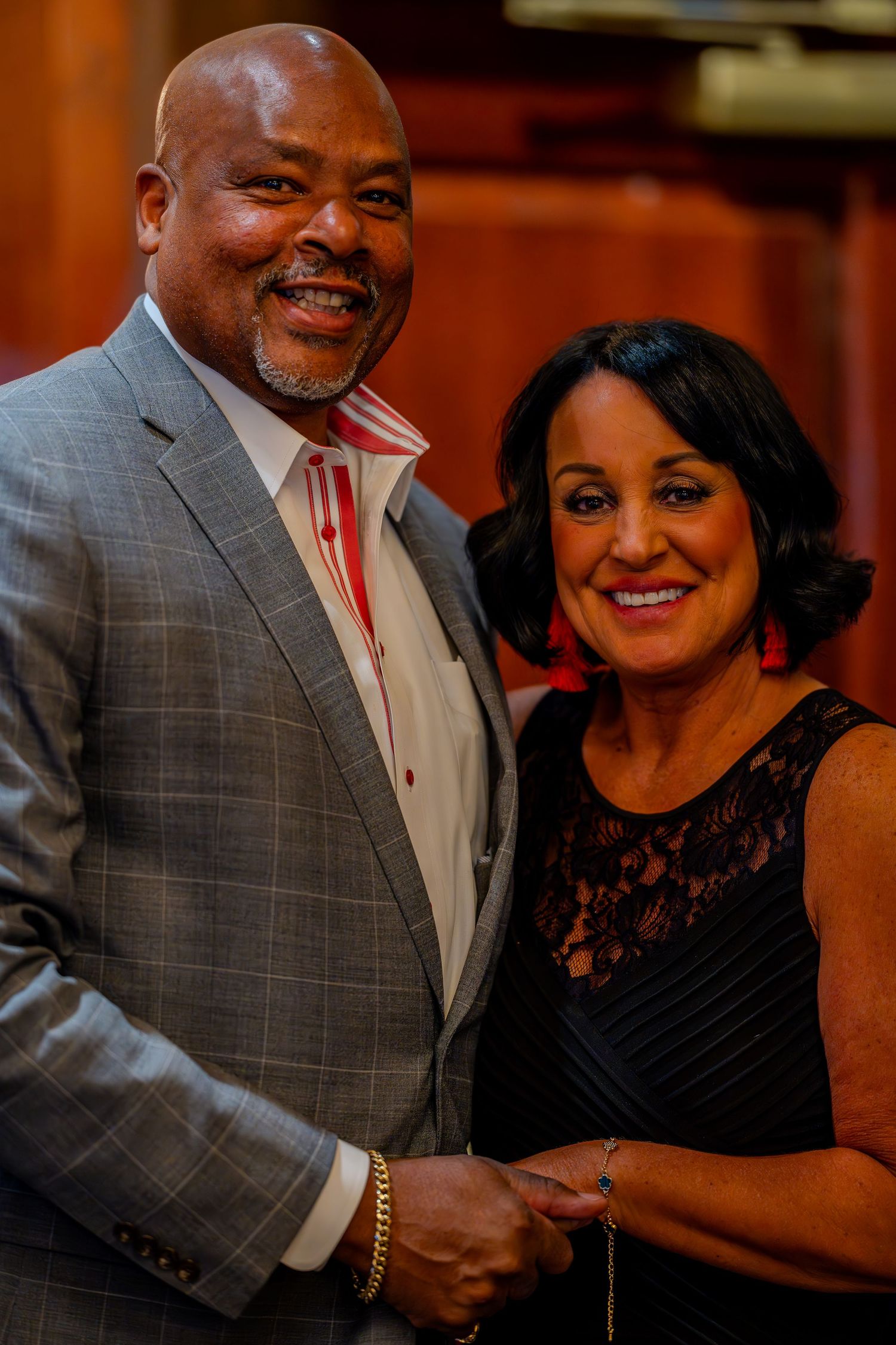 Smiling couple posing together at formal event, man in gray suit and woman in black dress with red earrings.