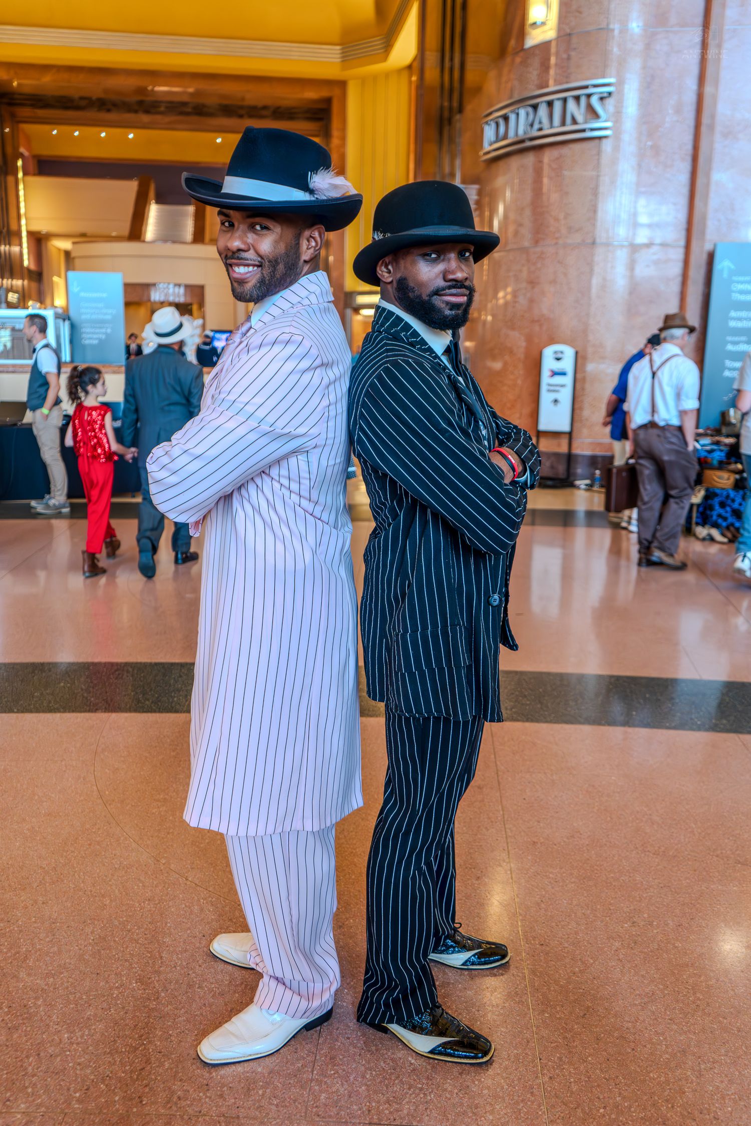 Two men in pinstripe zoot suits and fedora hats pose back-to-back with arms crossed in a grand hall.