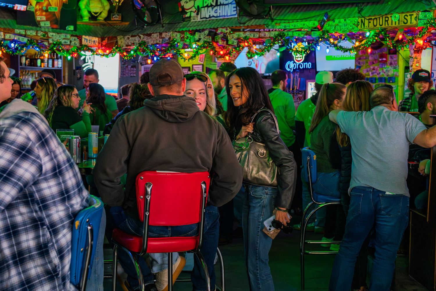 Crowded bar scene with colorful neon lights, patrons socializing and sitting on red stools in a festive indoor venue.