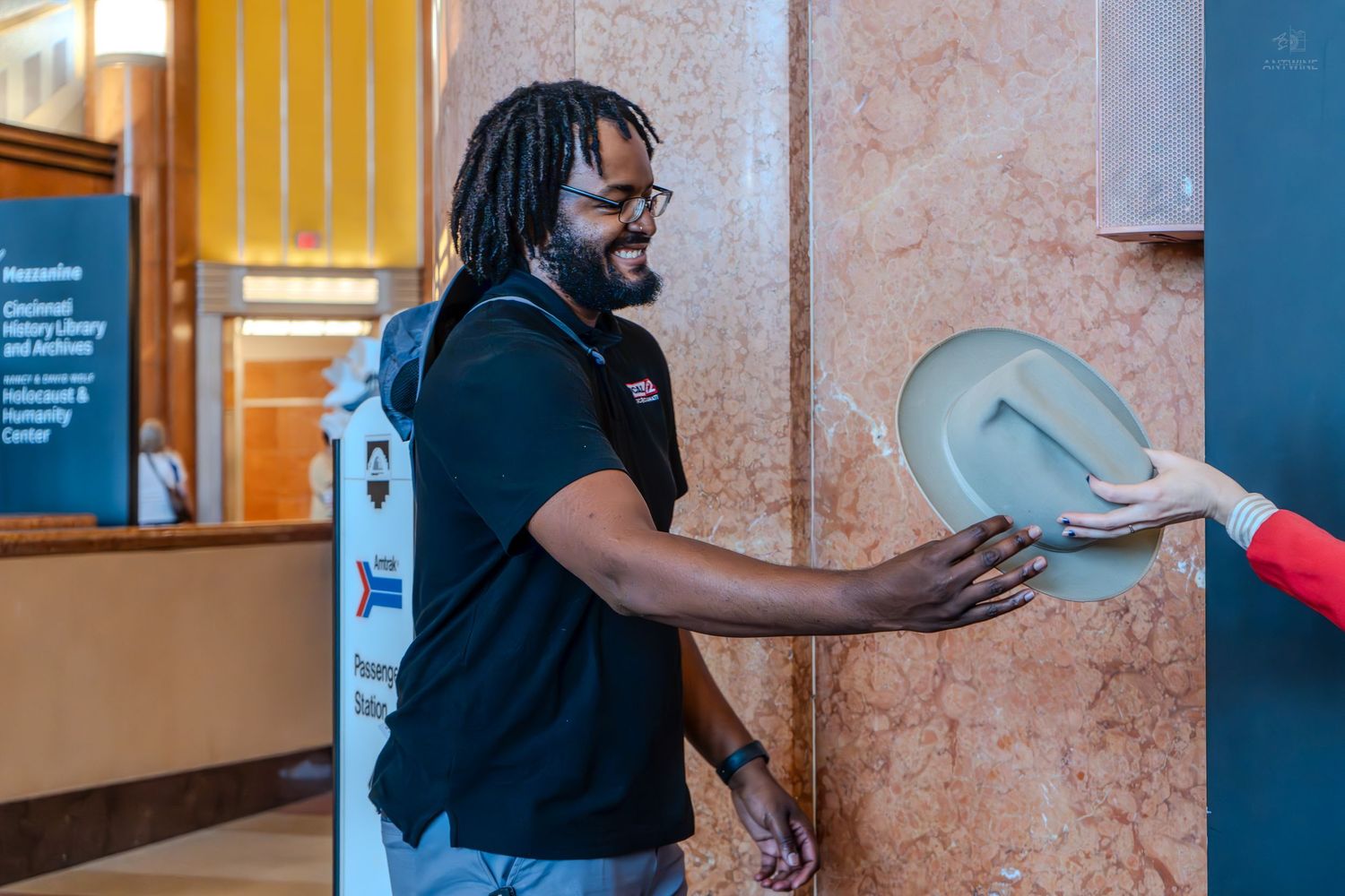 A smiling man in a black shirt receives a gray hat from someone in a marble lobby.