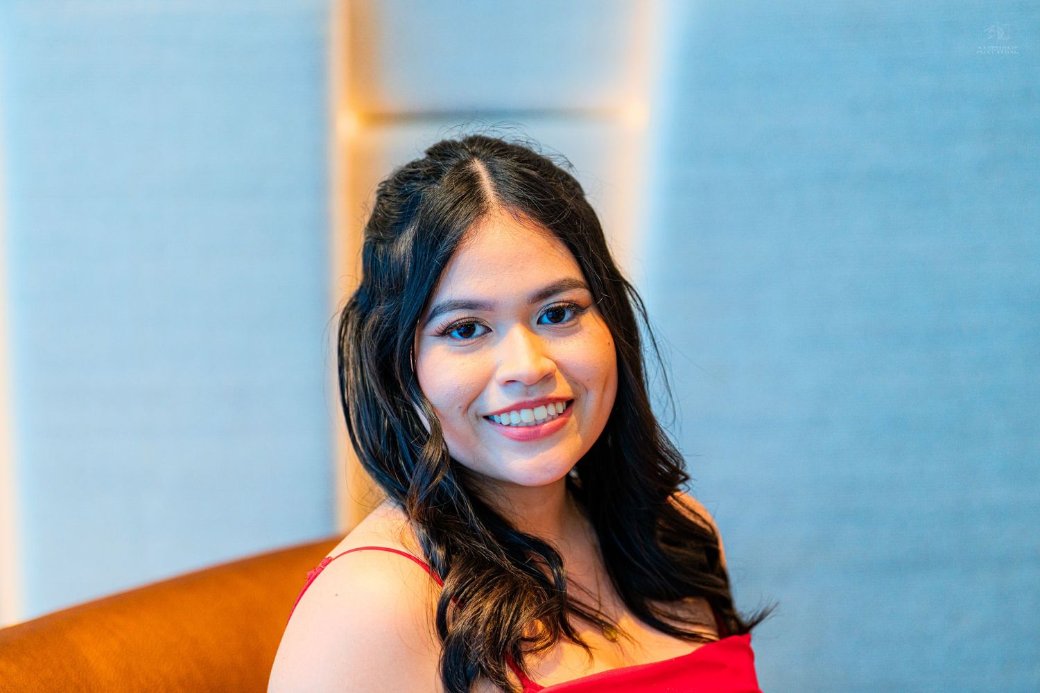 Smiling young woman with long dark hair wearing a red top, posed in a modern interior setting.