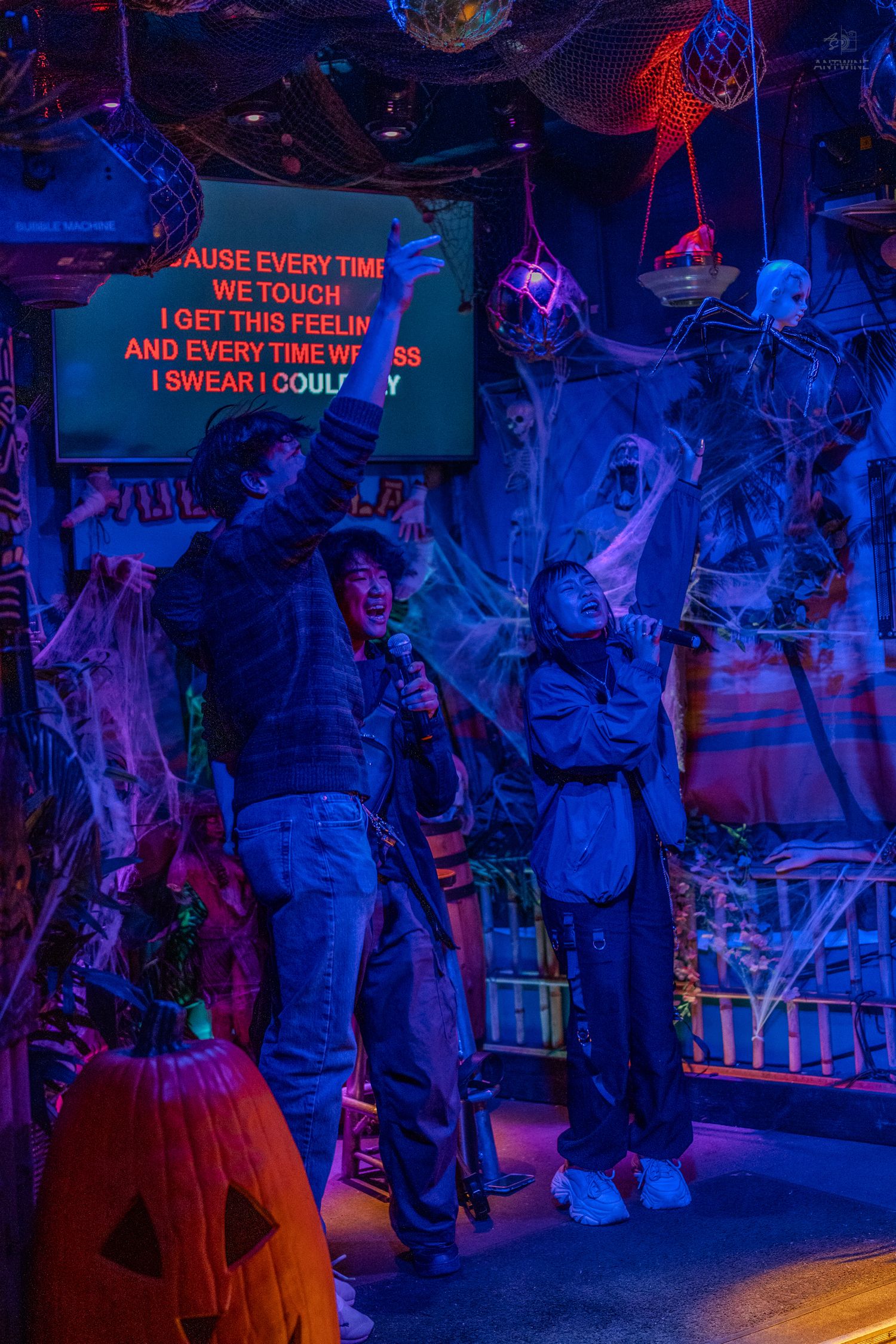 Two people singing karaoke together in a Halloween-themed bar with spooky decorations and neon lights.