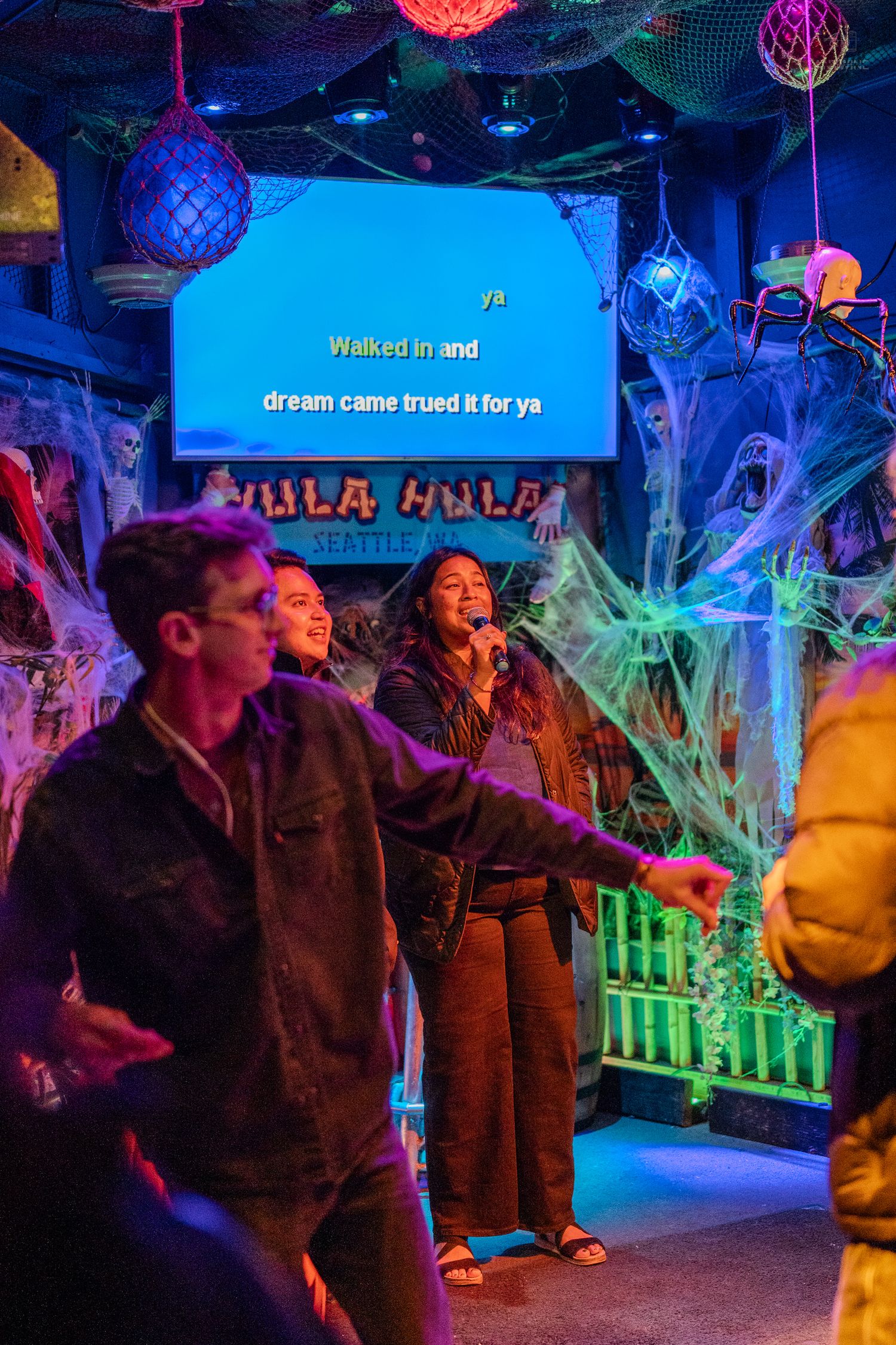 Group of friends enjoy karaoke night at Hula Hula bar surrounded by colorful Halloween decorations and neon lights.