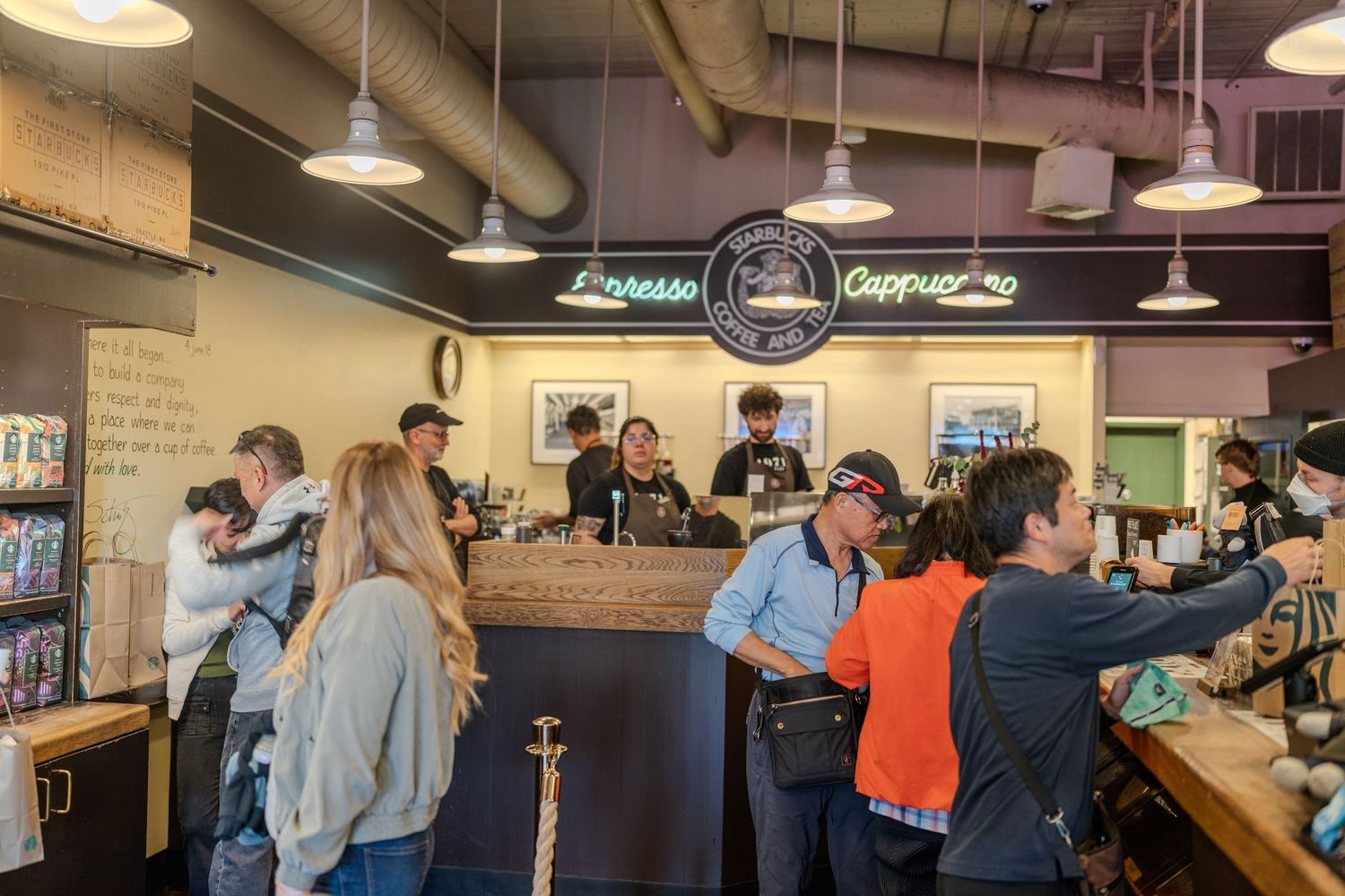 Busy coffee shop interior with customers at espresso and cappuccino bar under neon signs and pendant lights.