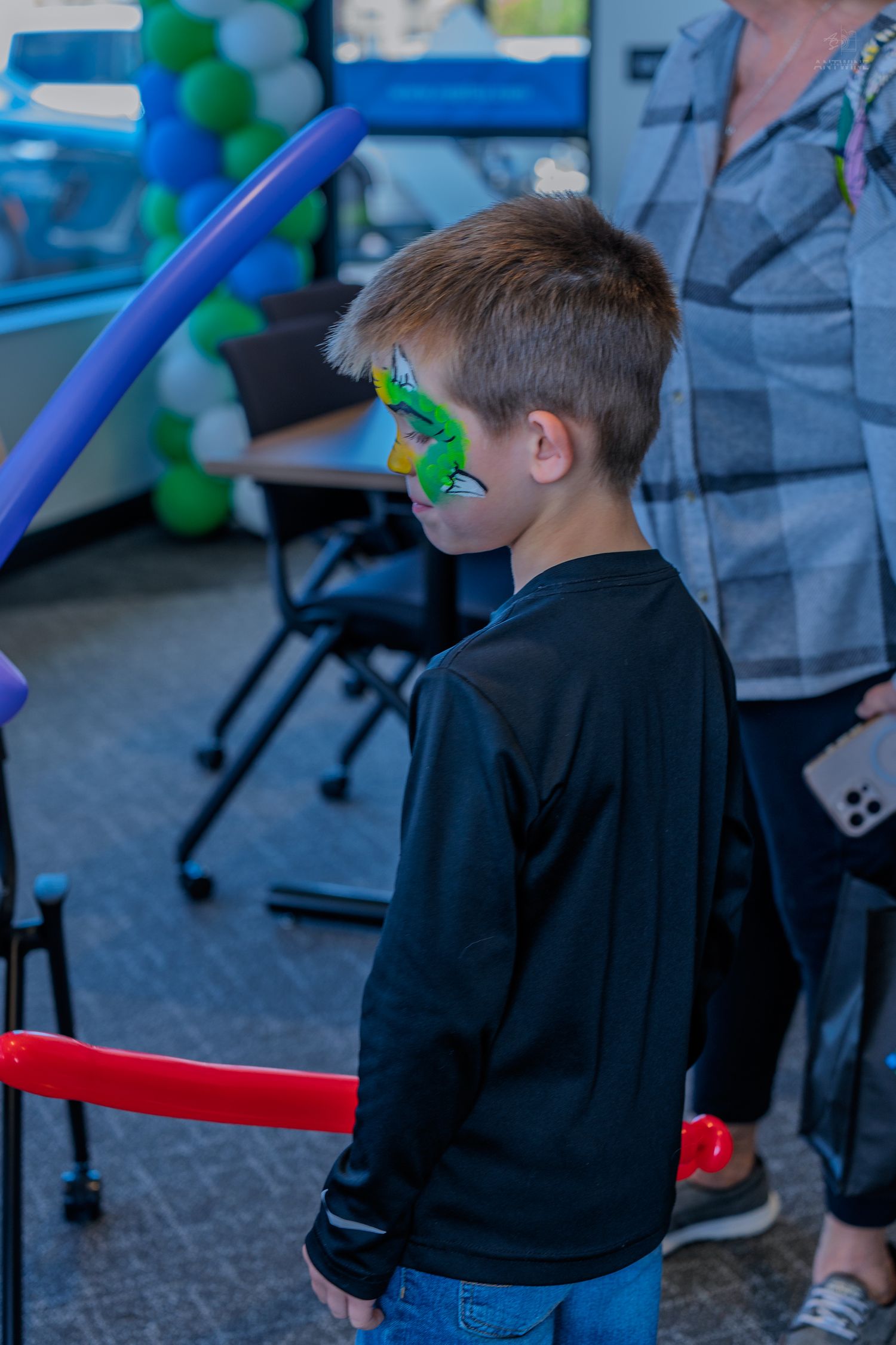 Boy with green dragon face paint wearing black shirt stands at a colorful balloon party event.