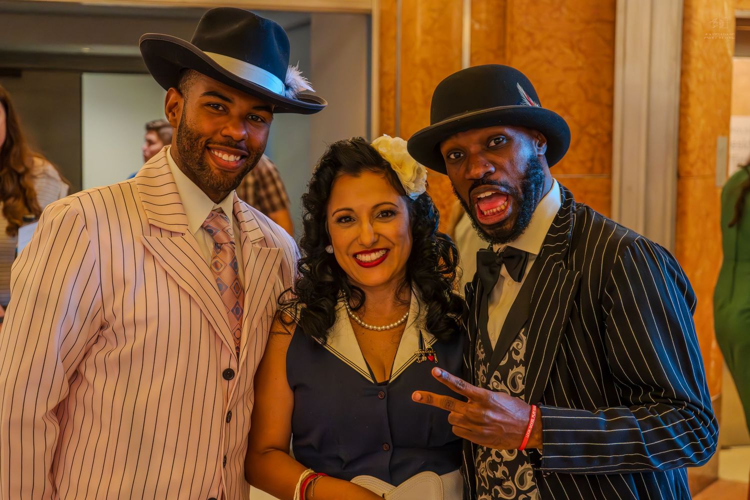 Three people in vintage 1920s-style outfits smile together at a formal event, wearing pinstripe suits and elegant dress.