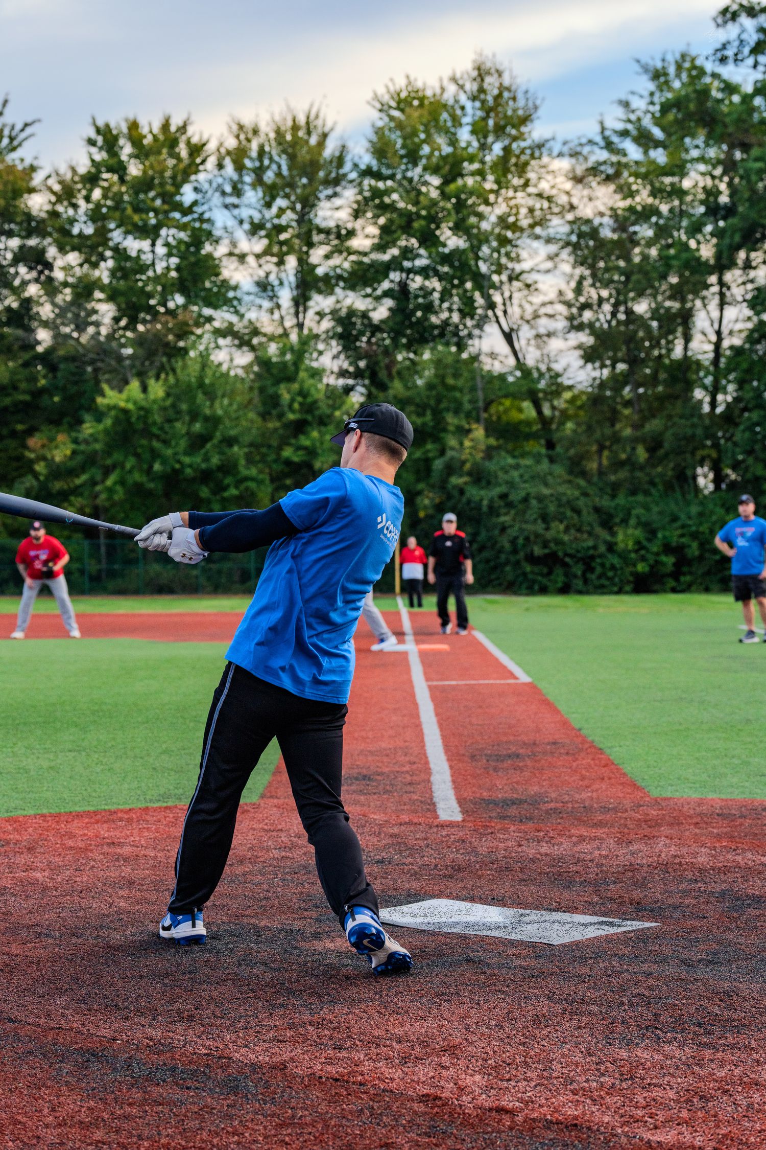 A batter in a blue uniform swings at home plate on a turf baseball field with players in the background.