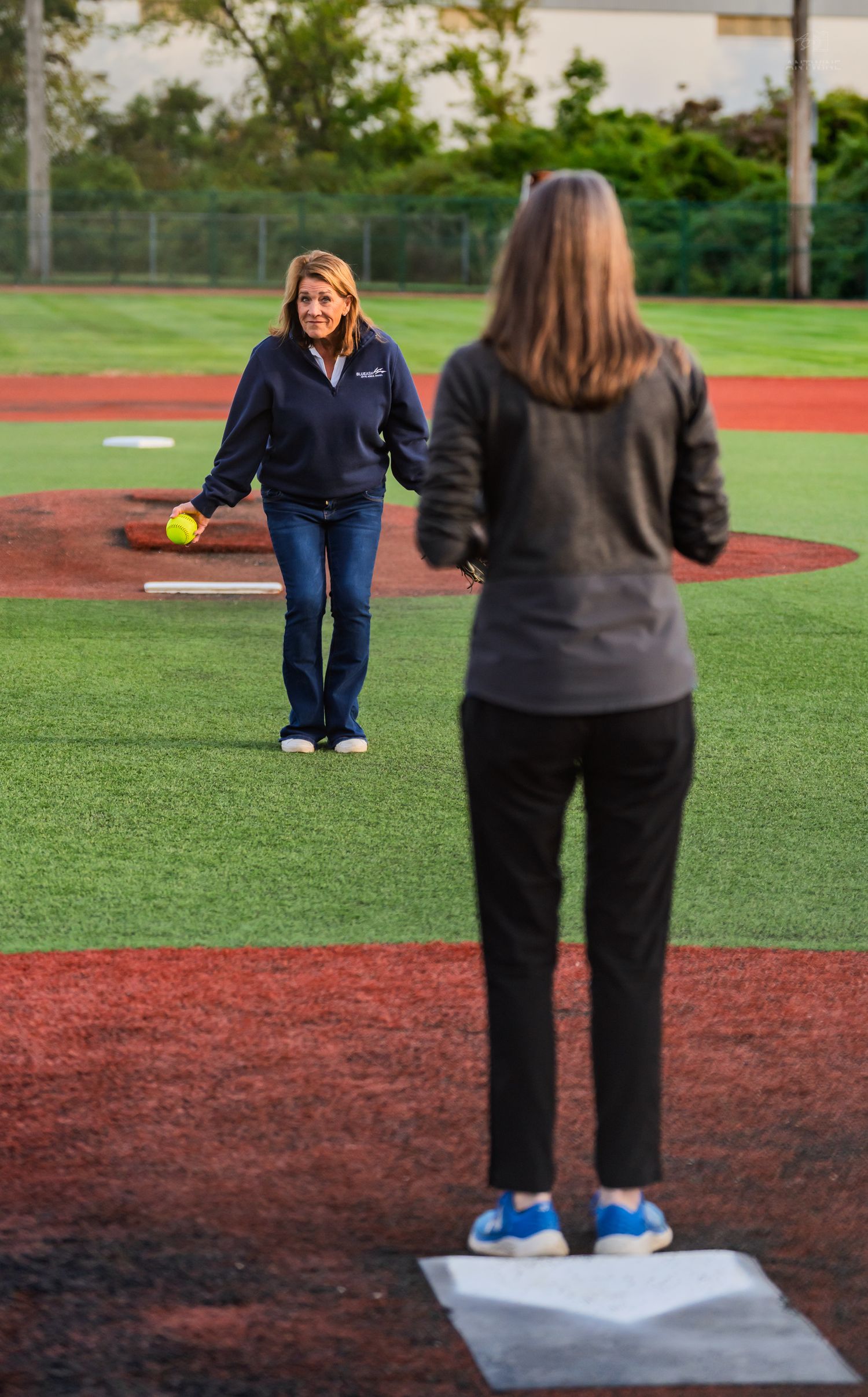 Woman prepares to throw yellow softball on pitcher's mound toward player standing at home plate outdoors.